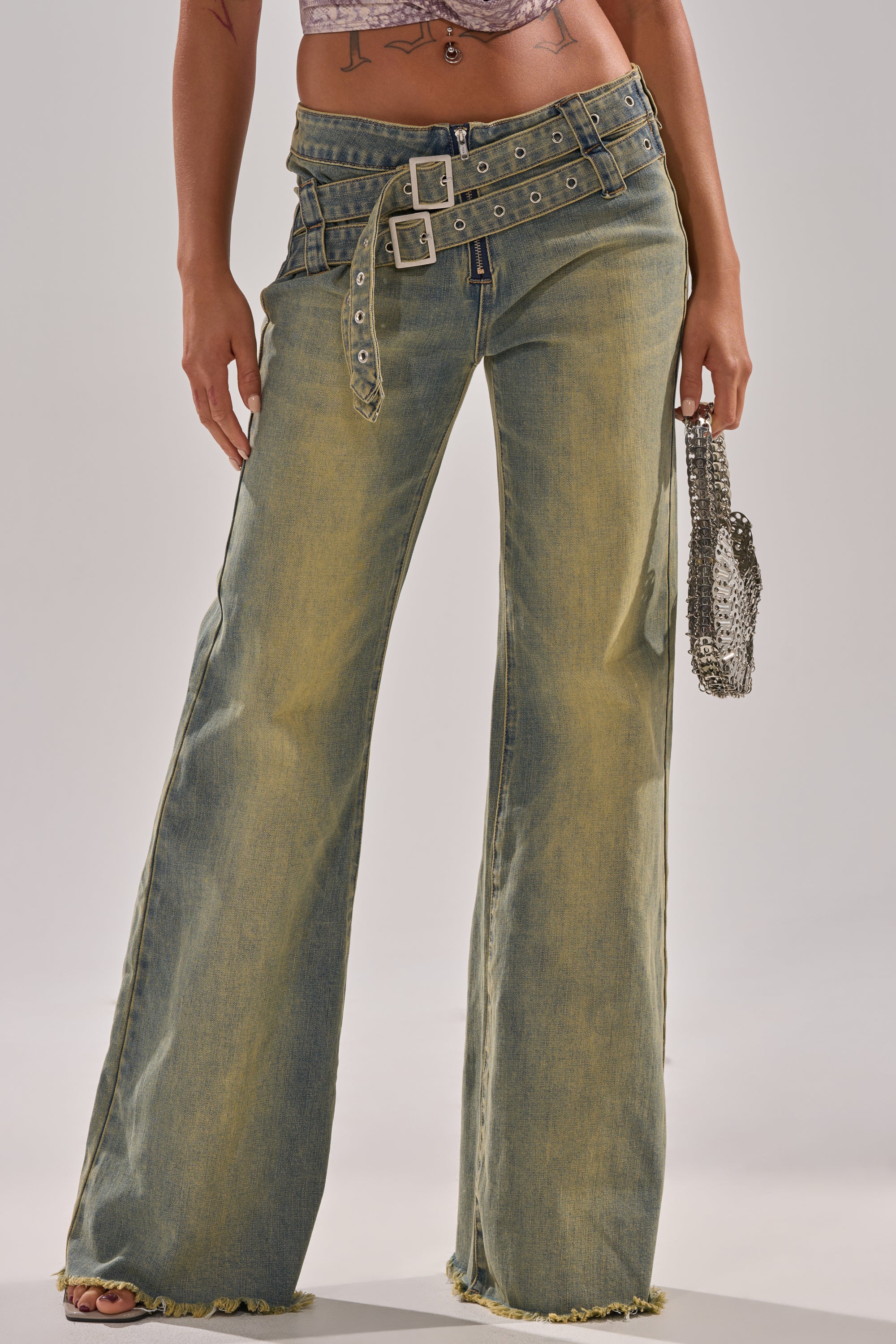 A person wears the JUST LIKE THAT STRAIGHT-LEG DENIM WITH BUCKLE, featuring a high waist, wide legs, distressed hems, and double-belt detail, and holds a small silver chain handbag—only their torso and legs are visible.