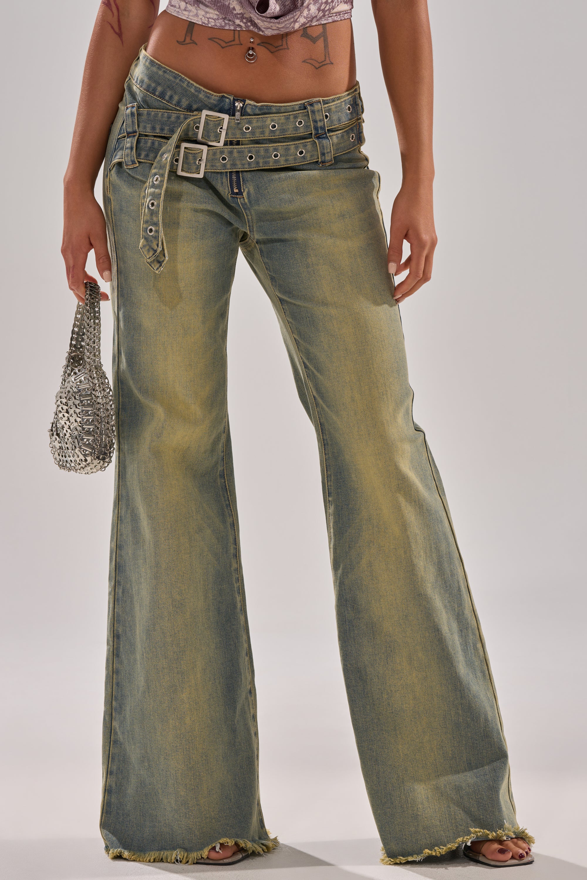 A person wears the JUST LIKE THAT STRAIGHT-LEG DENIM WITH BUCKLE—mid-rise, faded blue jeans with distressed hems and a double belt—paired with a small silver handbag, revealing their midriff and tattoos.