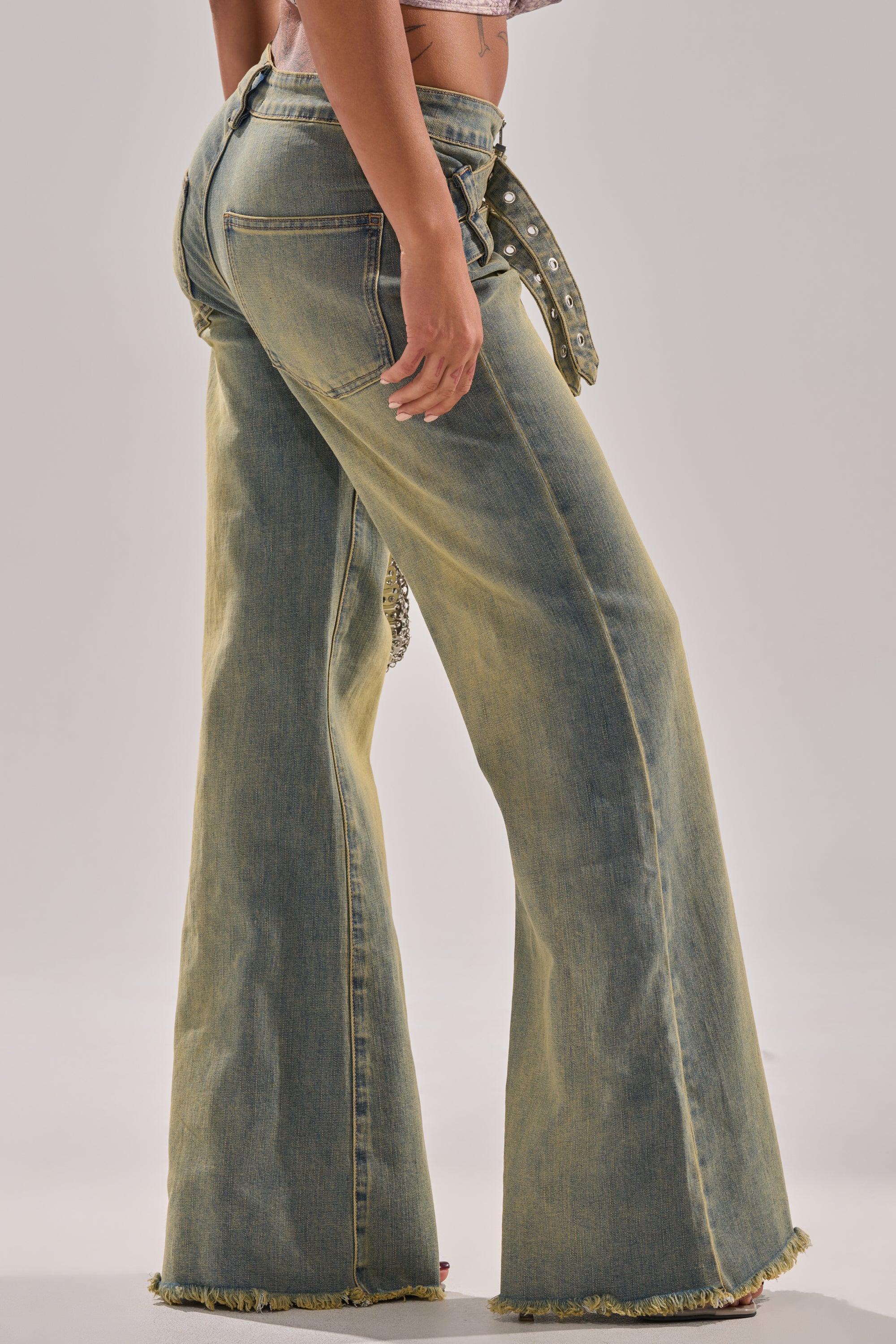 A person stands sideways in JUST LIKE THAT STRAIGHT-LEG DENIM WITH BUCKLE—high-waisted, wide-leg, light-wash jeans with a distressed hem and a studded buckle belt—against a plain, neutral background.
