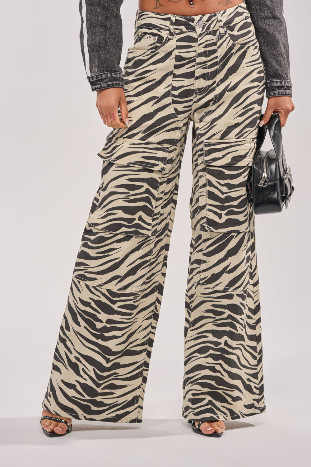 A person wears IN THE WILD ZEBRA DENIM high-waisted, wide-leg pants with black open-toe heels, a dark cropped jacket, and holds a small black handbag against a plain white background. 0