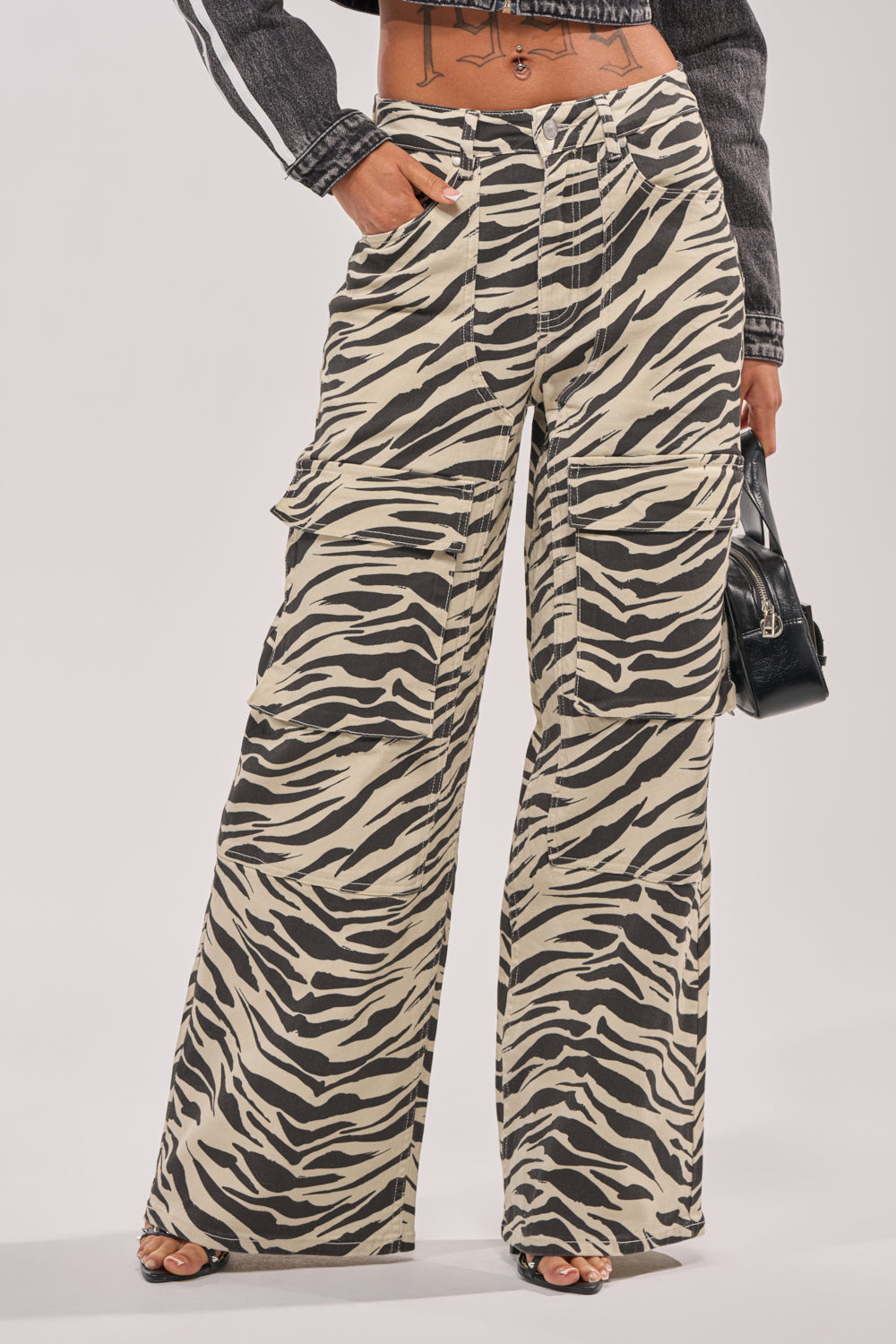 A person wearing the IN THE WILD ZEBRA DENIM high-waisted, wide-leg pants, paired with a cropped black denim jacket and carrying a small black handbag. 1