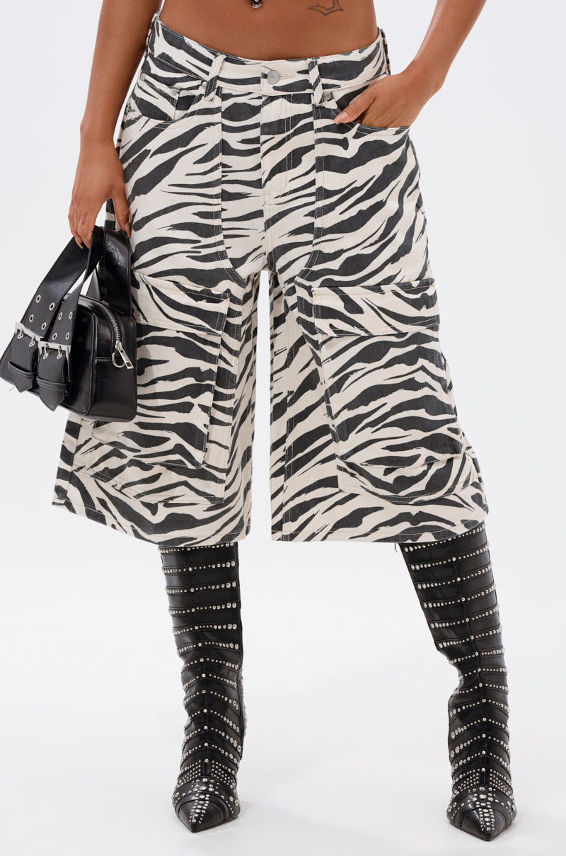 Wearing the ZEBRA LUXE BERMUDA SHORT, black studded knee-high boots, and holding a small black handbag, a person stands against a white background.