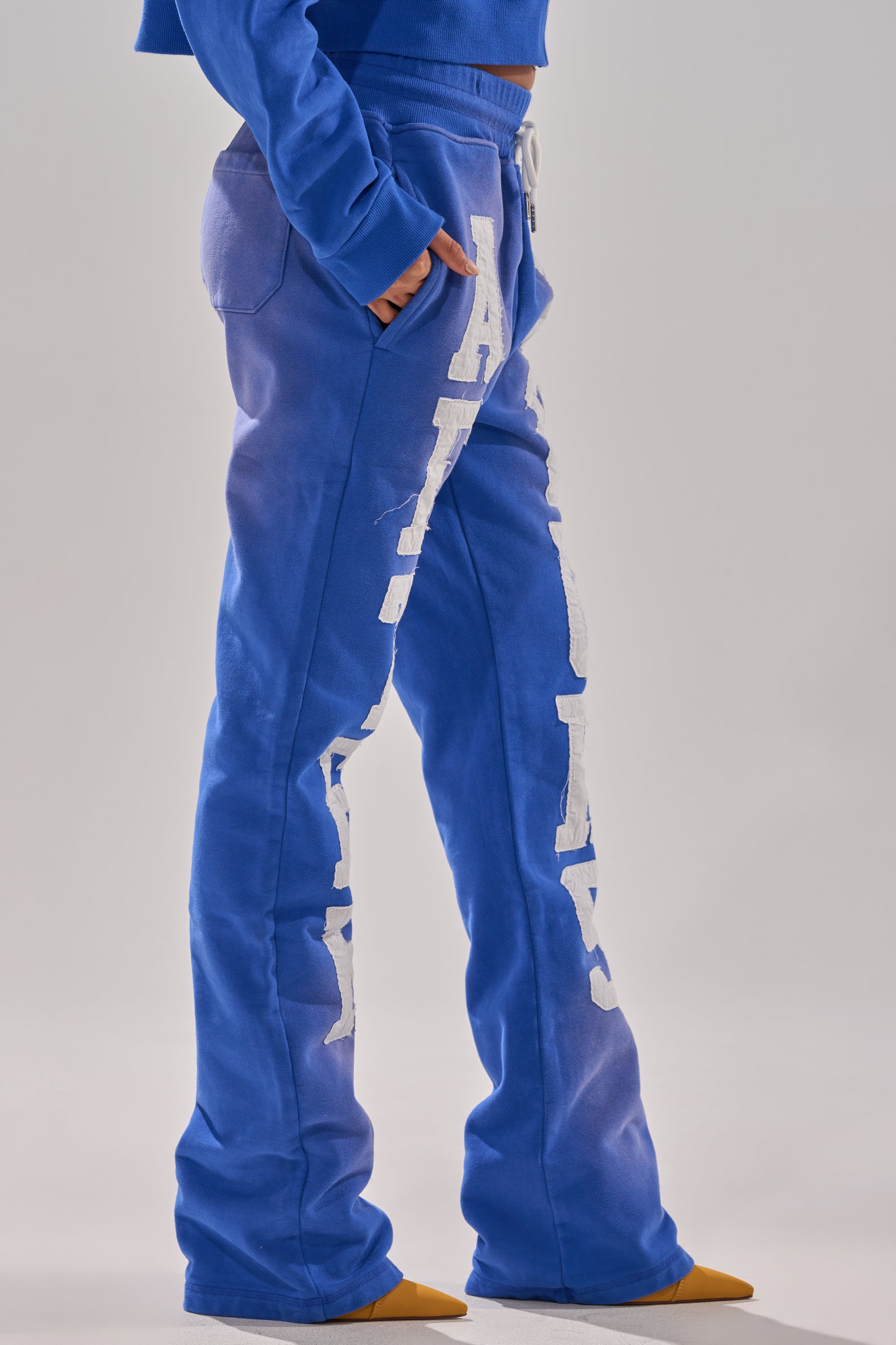 Person in AFTER HOURS FLARED SWEATPANT, featuring blue high-rise cotton fabric with bold white side lettering and a relaxed flared fit that slightly covers orange-toed shoes, paired with a matching blue top. 2