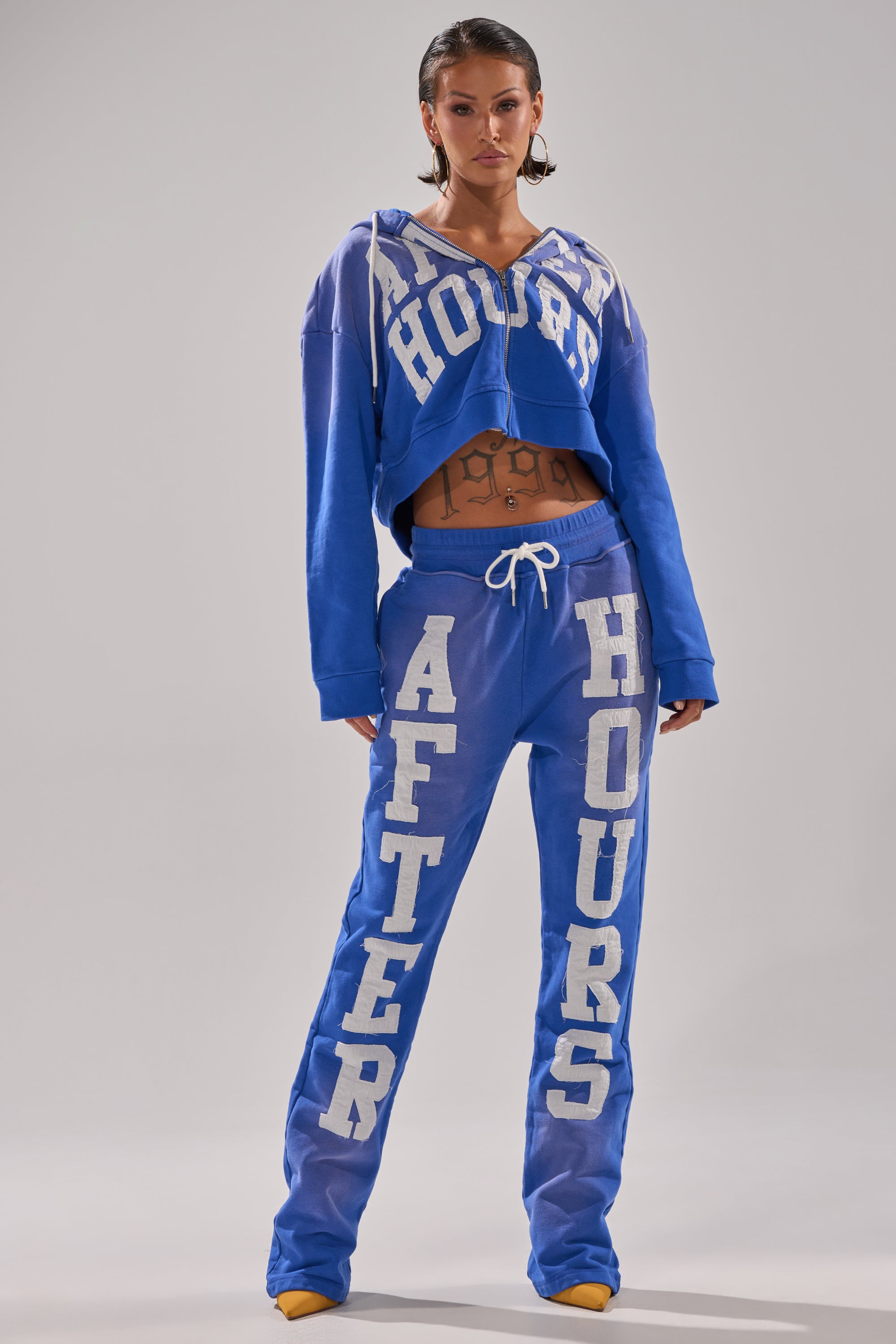 A woman faces forward in blue AFTER HOURS FLARED SWEATPANTS with bold white lettering, paired with hoop earrings and yellow pointed shoes, posing against a plain gray background. 4