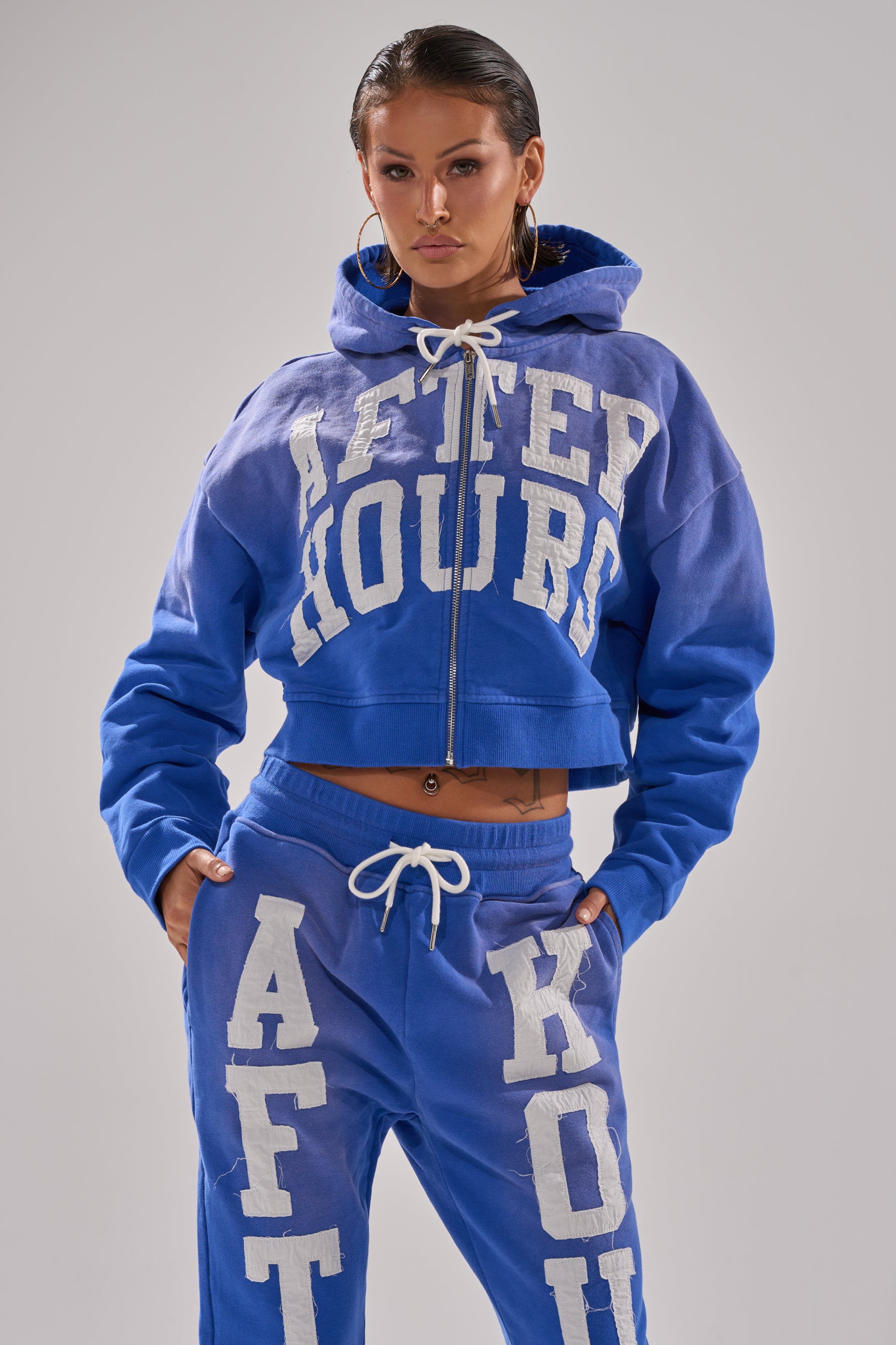 A person wearing the AFTER HOURS ZIP UP SWEATSHIRT in bright blue with bold white "AFTER HOURS" lettering and matching sweatpants, posing with hands in pockets against a plain background.
