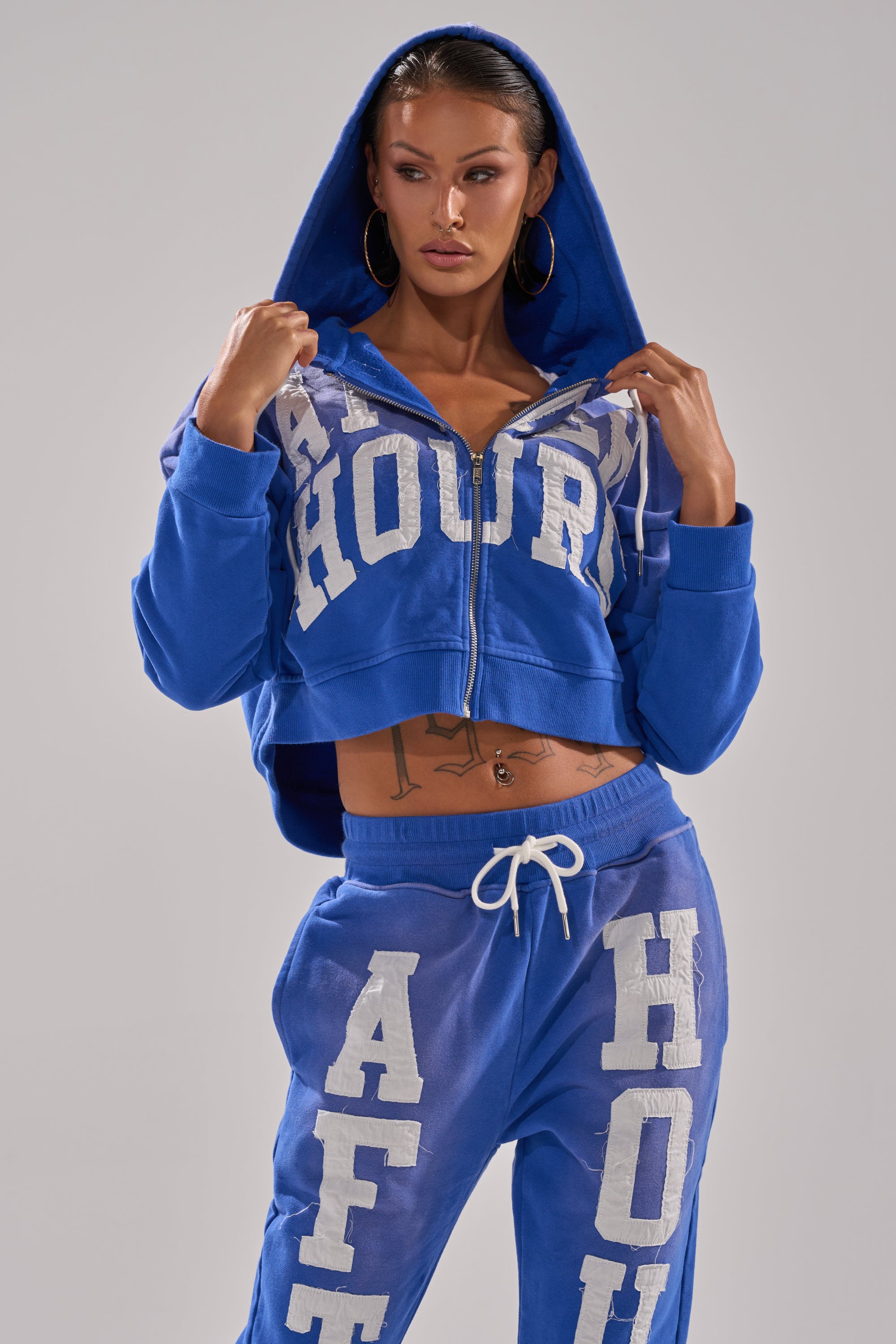 A woman wears the AFTER HOURS ZIP UP SWEATSHIRT in blue with bold white lettering, hood up and unzipped, revealing tattoos on her midriff, paired with matching sweatpants.