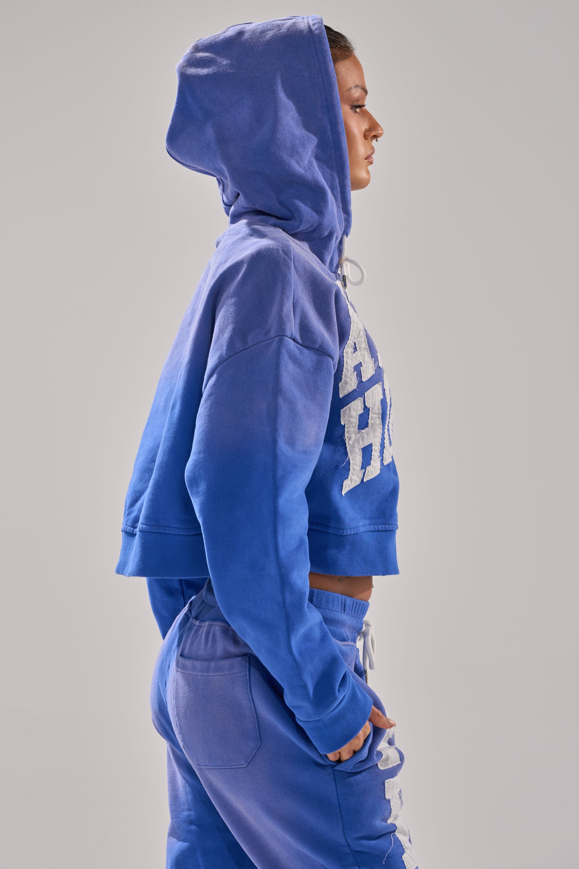 A person wearing the AFTER HOURS ZIP UP SWEATSHIRT, an oversized blue hooded cotton sweatshirt, with matching blue sweatpants stands in profile against a plain light gray background.