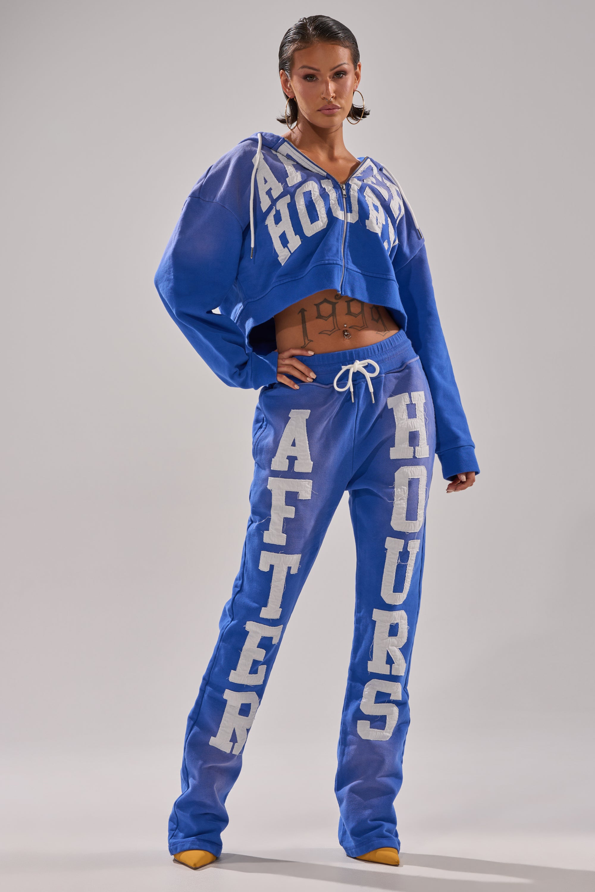 A woman with slicked-back hair and torso tattoos wears the AFTER HOURS ZIP UP SWEATSHIRT and matching blue sweatpants, featuring bold white "AFTER HOURS" lettering. She completes the look with yellow shoes.