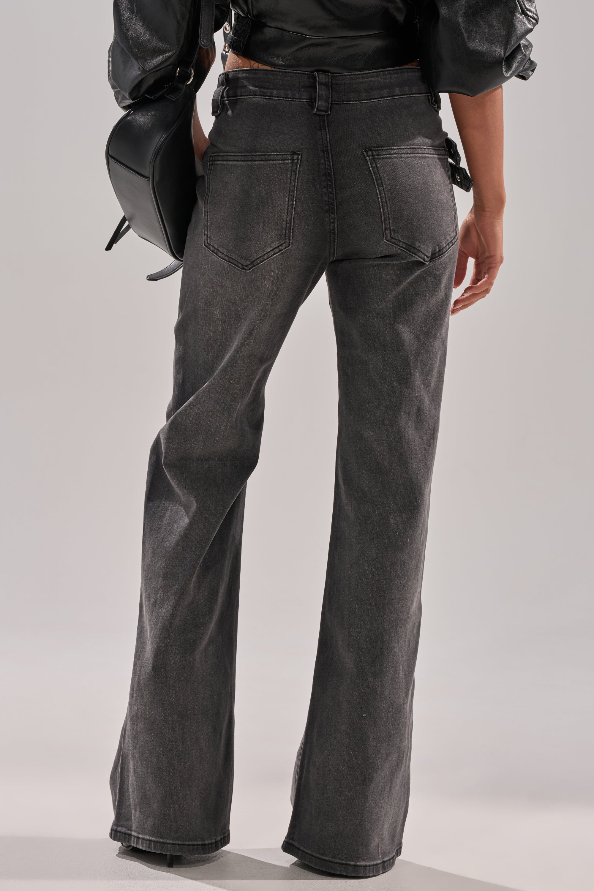 A person stands with their back to the camera, wearing WONT LOSE MYSELF WIDE LEG DENIM high-waisted, washed-black jeans with an adjustable waistbelt and holding a black handbag. 4