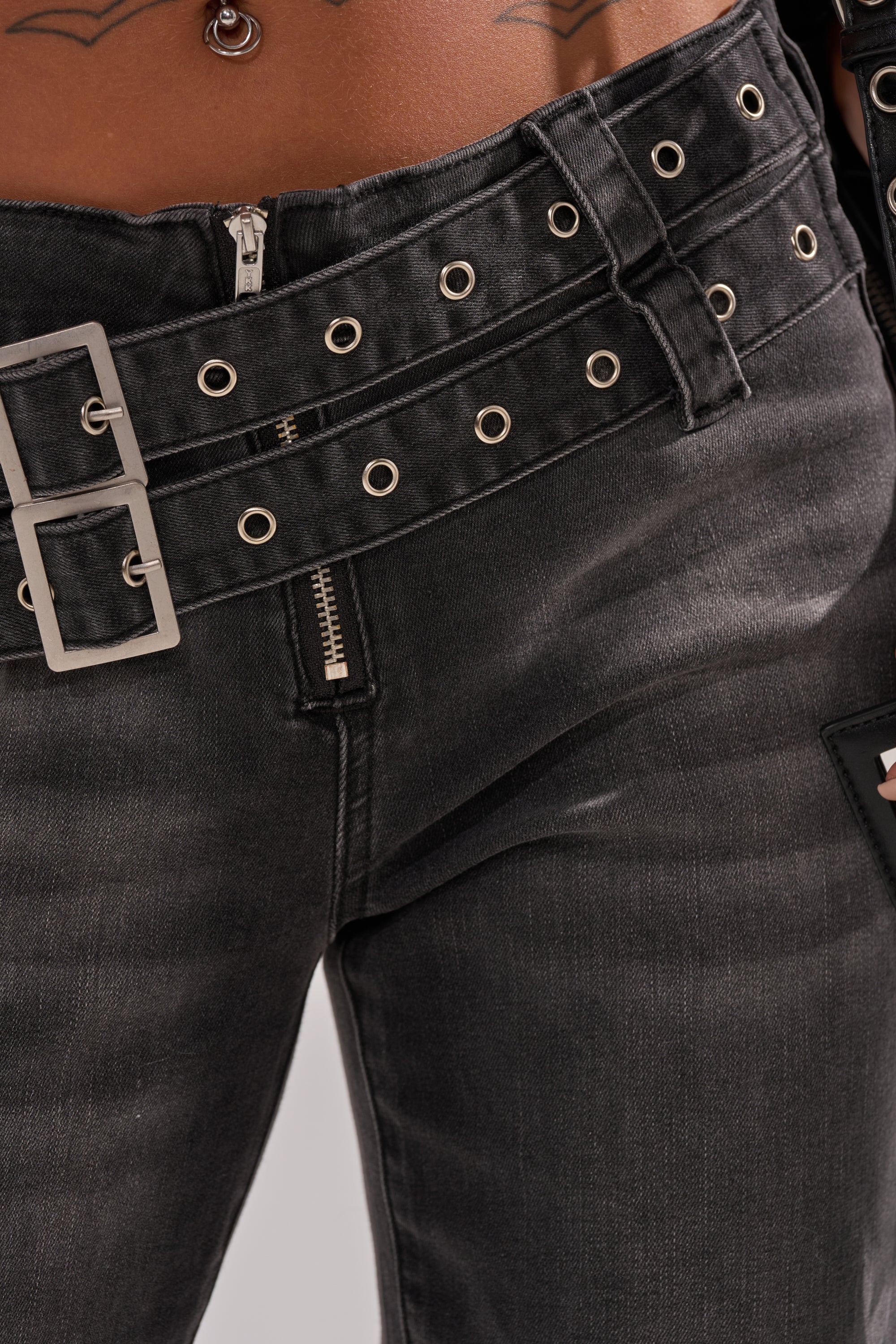 Close-up of a person wearing WONT LOSE MYSELF WIDE LEG DENIM in washed black, styled with two silver-buckled, grommeted belts, partially unzipped fly, plus visible navel piercing and tattoos above the waistband. 7