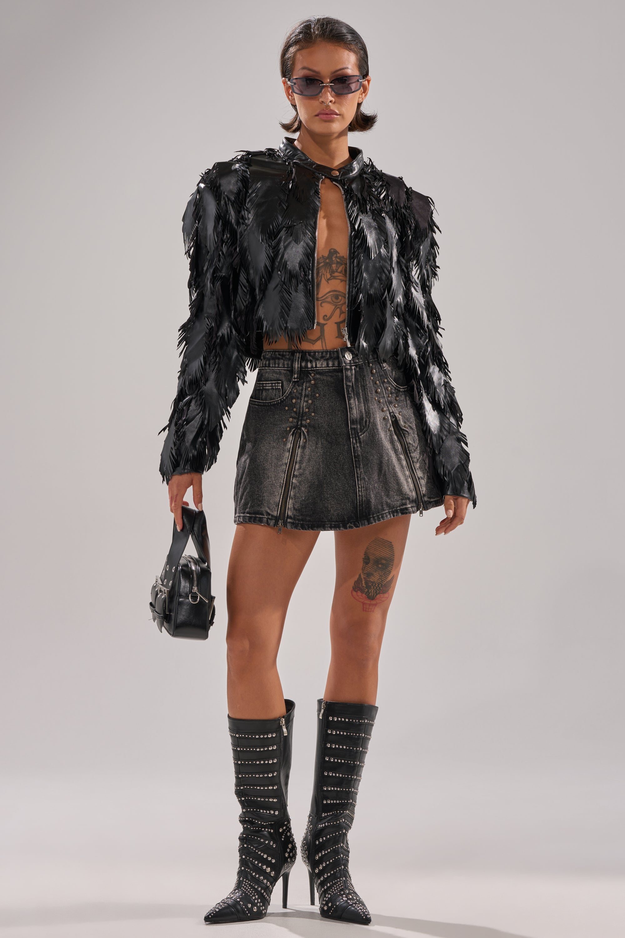 A woman confidently poses against a plain backdrop wearing sunglasses, a black feathered jacket, black studded knee-high boots, a black handbag, and the GET IT GIRL DENIM MINI SKIRT. Tattoos are visible on her torso and leg.