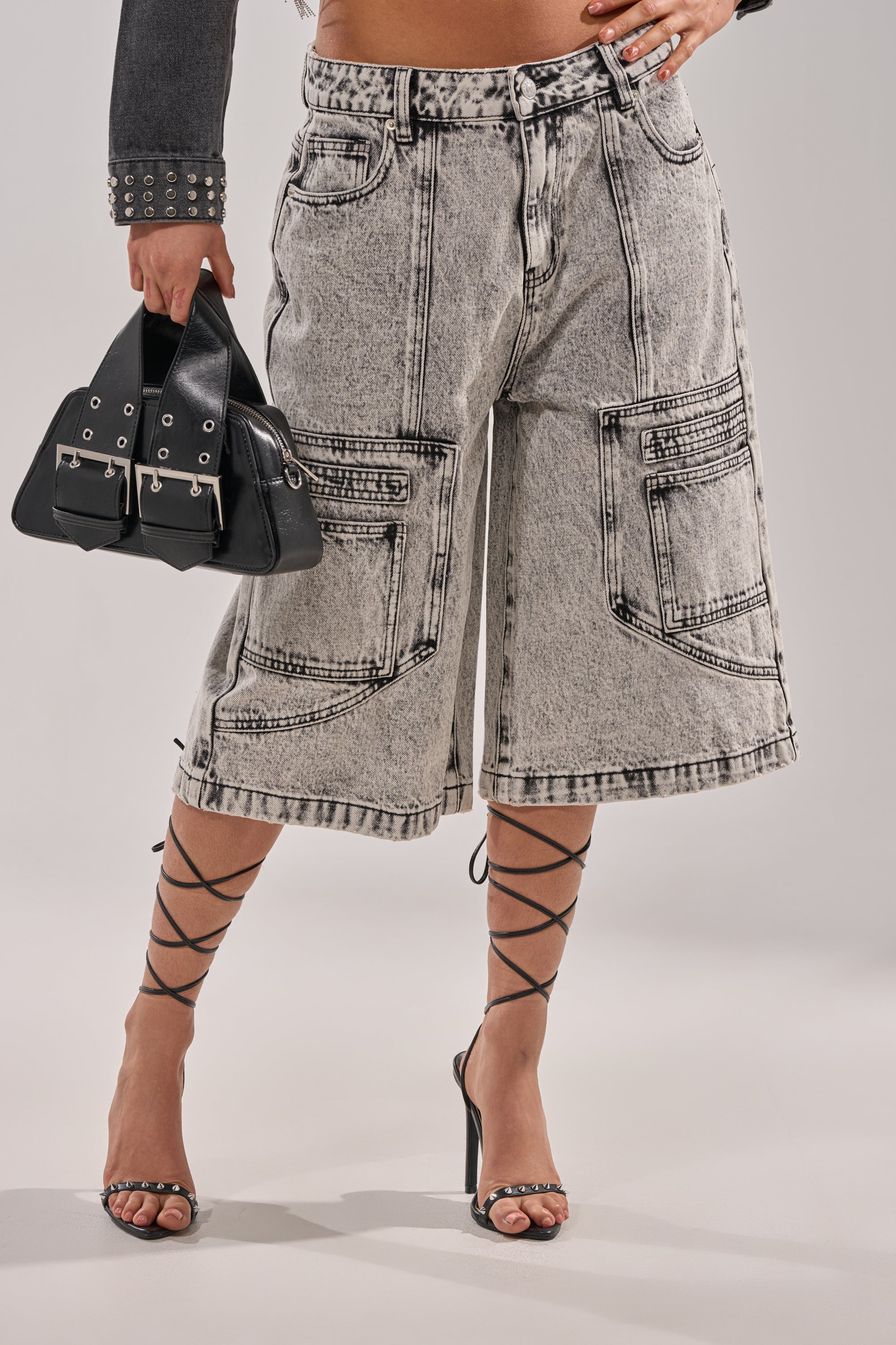 The lower half of a person wearing TROUBLE MAKER BERMUDA SHORT high-rise acid-wash denim shorts, black strappy high-heeled sandals, and holding a small black buckle handbag is visible against a plain background. 2