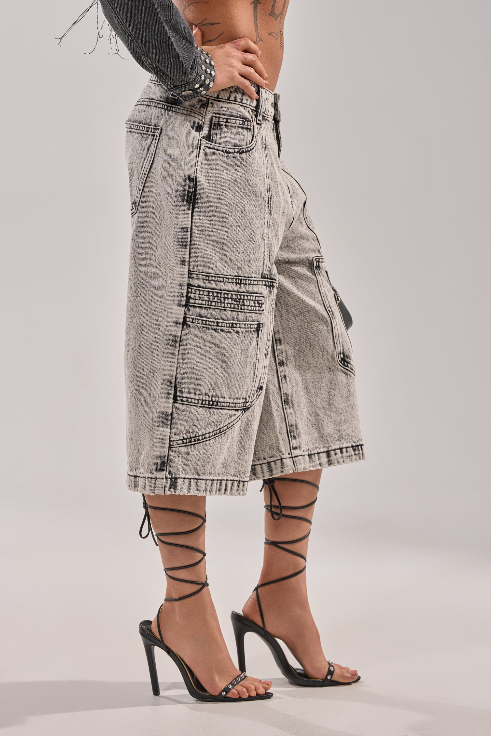 A person wearing TROUBLE MAKER BERMUDA SHORT—light-wash, high-rise denim bermuda shorts with cargo pockets—and black strappy high-heeled sandals, standing against a plain background. 3