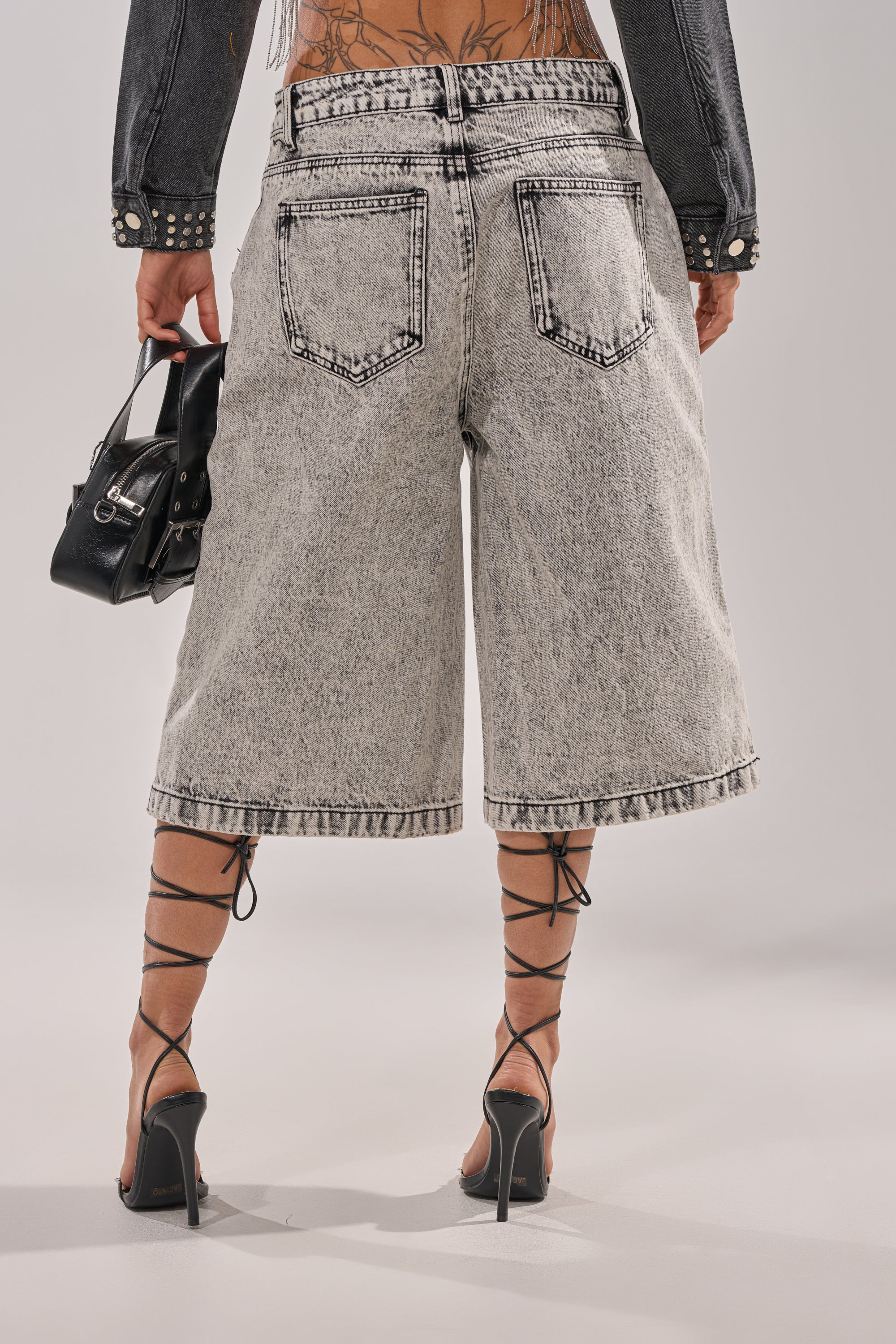 Wearing TROUBLE MAKER BERMUDA SHORT high-rise acid-wash denim bermuda shorts, strappy black heels, and holding a small black handbag, a person shows off a lower back tattoo. 4