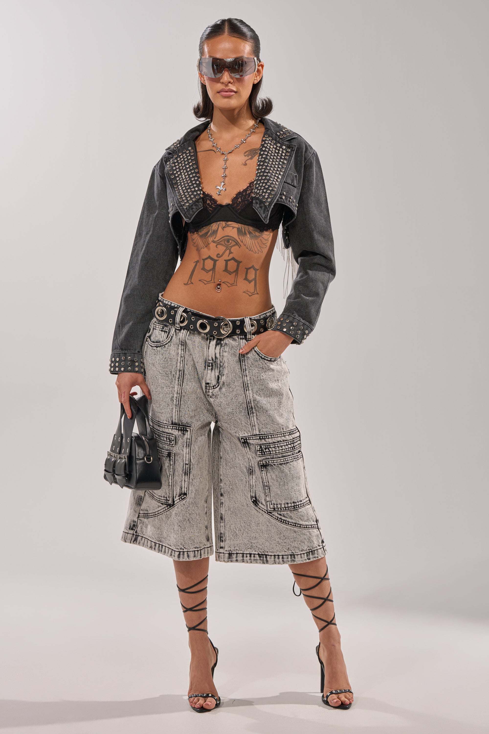 A woman wearing large sunglasses, a studded cropped denim jacket, black bralette, TROUBLE MAKER BERMUDA SHORT with cargo pockets, lace-up heels, holding a small black handbag, and showing torso tattoos. 6