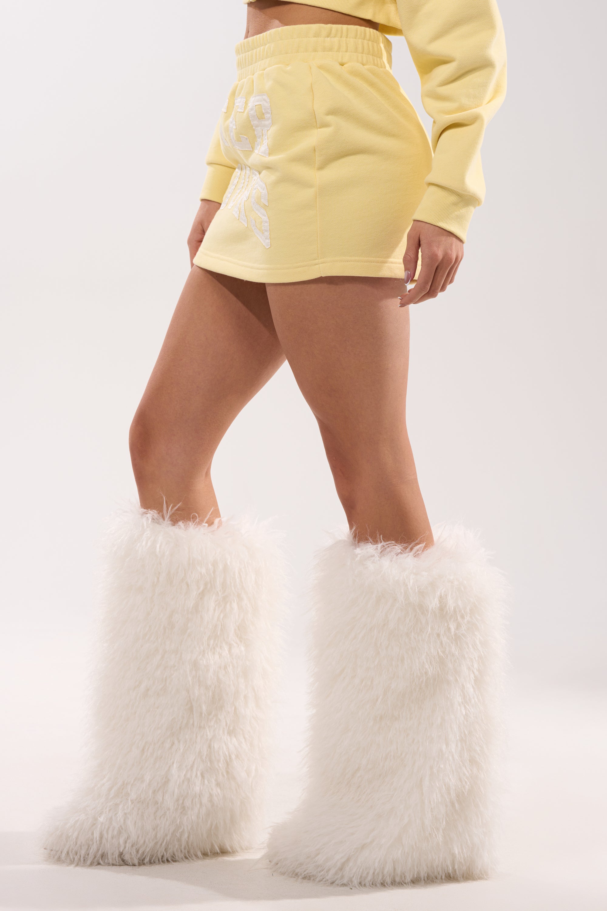 A person wears the AFTER HOURS MINI SKIRT IN YELLOW with a matching pastel top and large white fluffy knee-high boots, set against a plain light background that highlights the skirt and standout boots. 4