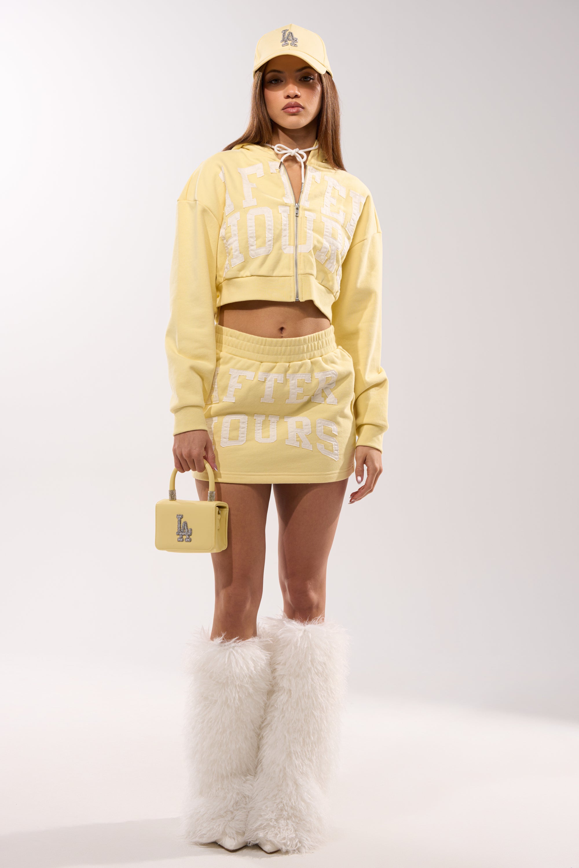 A woman wears a pastel yellow cropped hoodie and high-rise AFTER HOURS MINI SKIRT IN YELLOW, paired with a yellow cap, small yellow handbag, and tall white furry boots, standing against a plain light background. 6