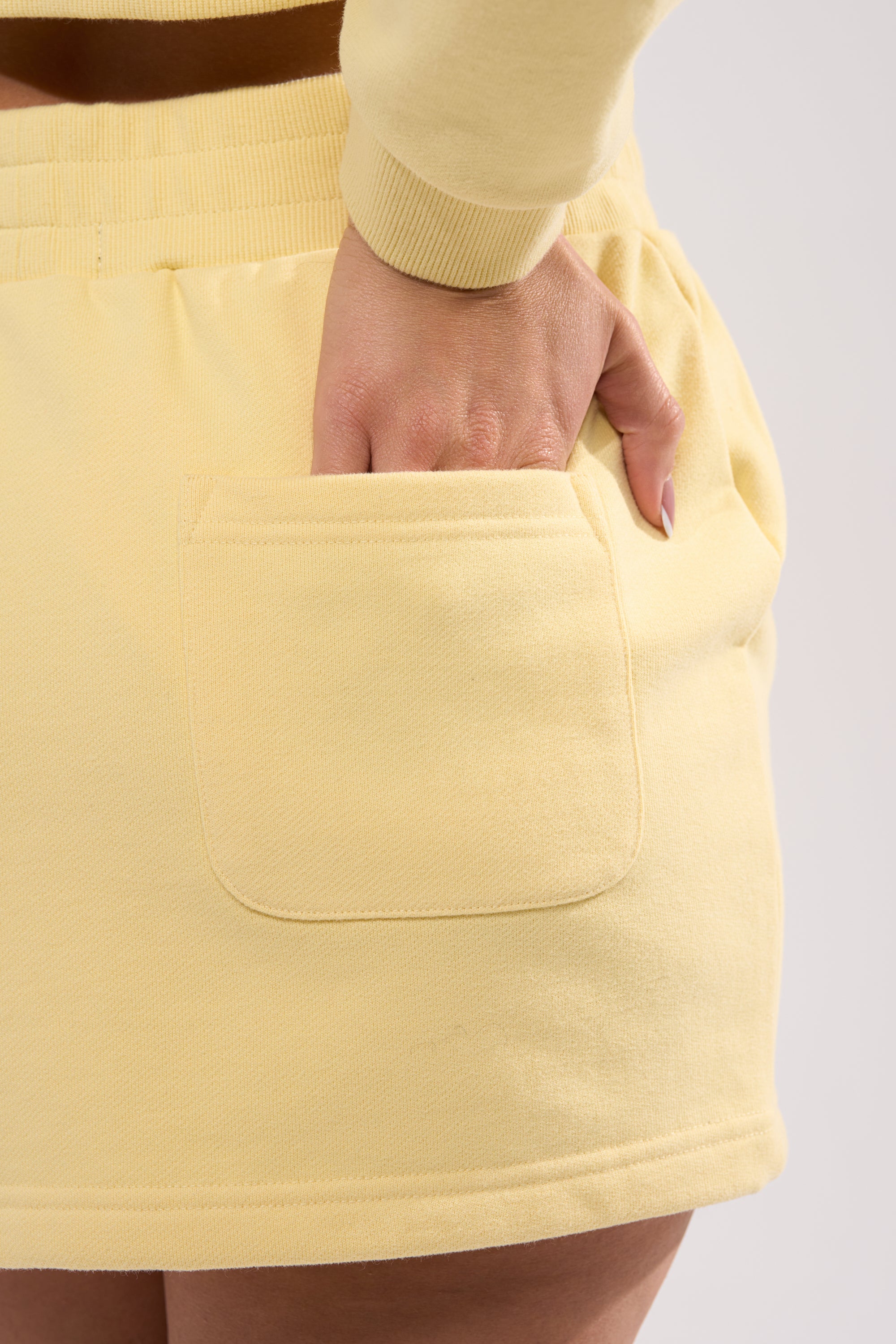 A person wearing the AFTER HOURS MINI SKIRT IN YELLOW with a matching long-sleeve top has their right hand in the skirt's front pocket. The background is plain and light-colored. 8