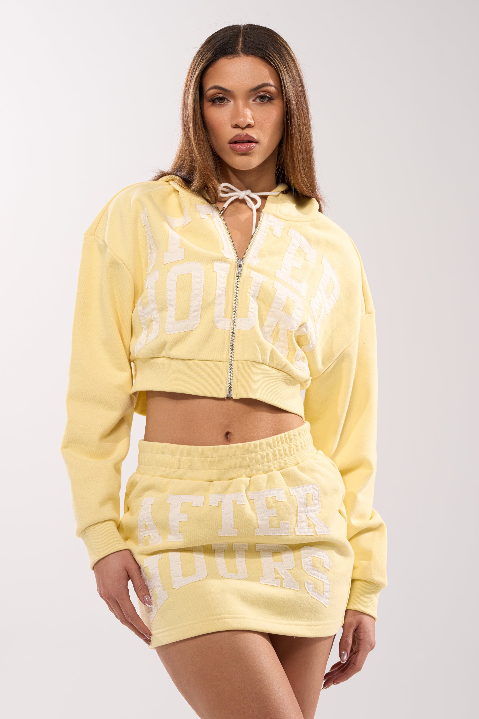 A woman models the AFTER HOURS SWEATSHIRT IN YELLOW—a butter yellow cropped sweatshirt with "AFTER HOURS" in bold white letters—paired with a matching high-waisted mini skirt against a plain light background. 0