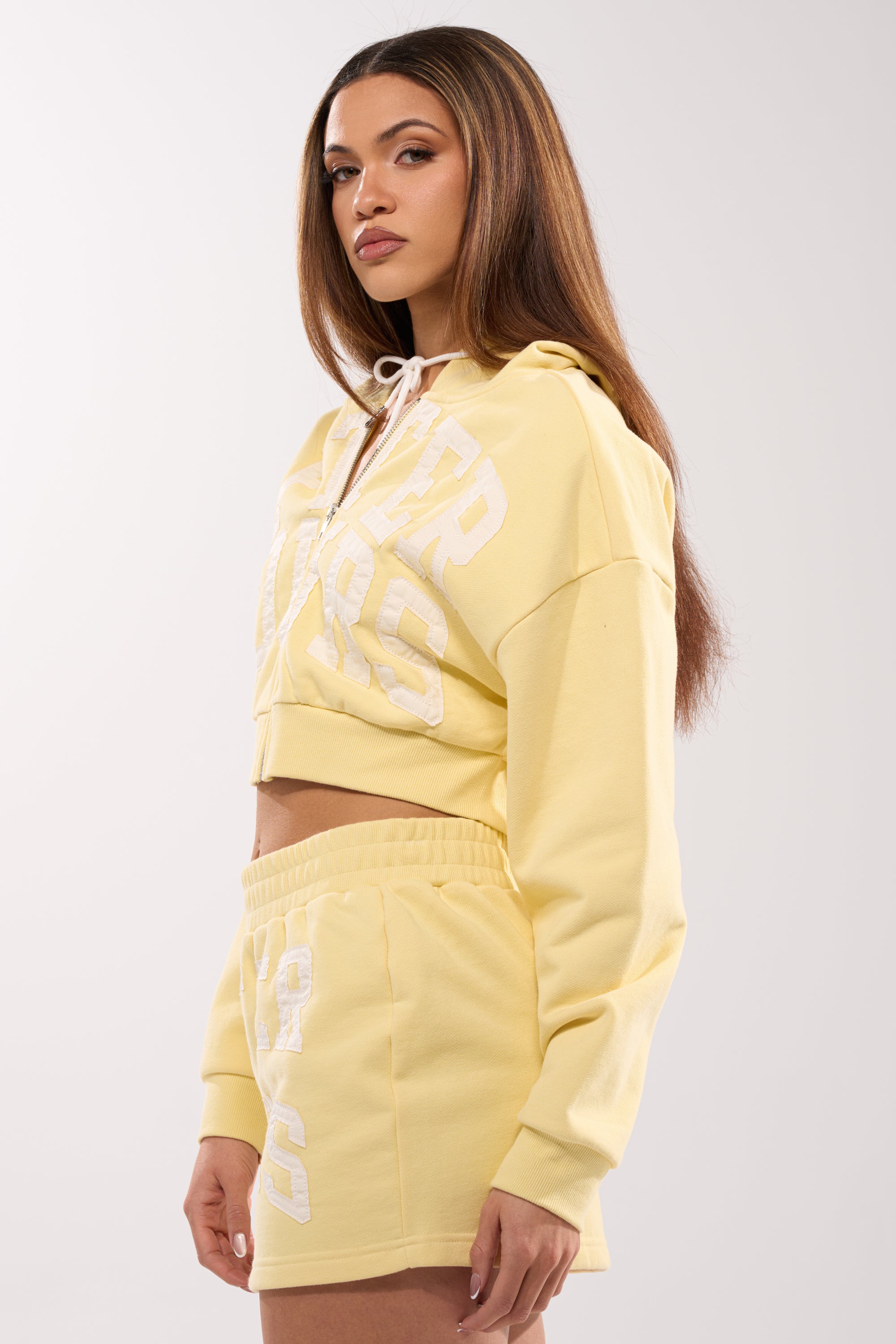 A woman with long straight hair models the AFTER HOURS SWEATSHIRT IN YELLOW, a butter yellow cropped top with bold white lettering and matching shorts, posing against a plain light background and gazing to the side. 4