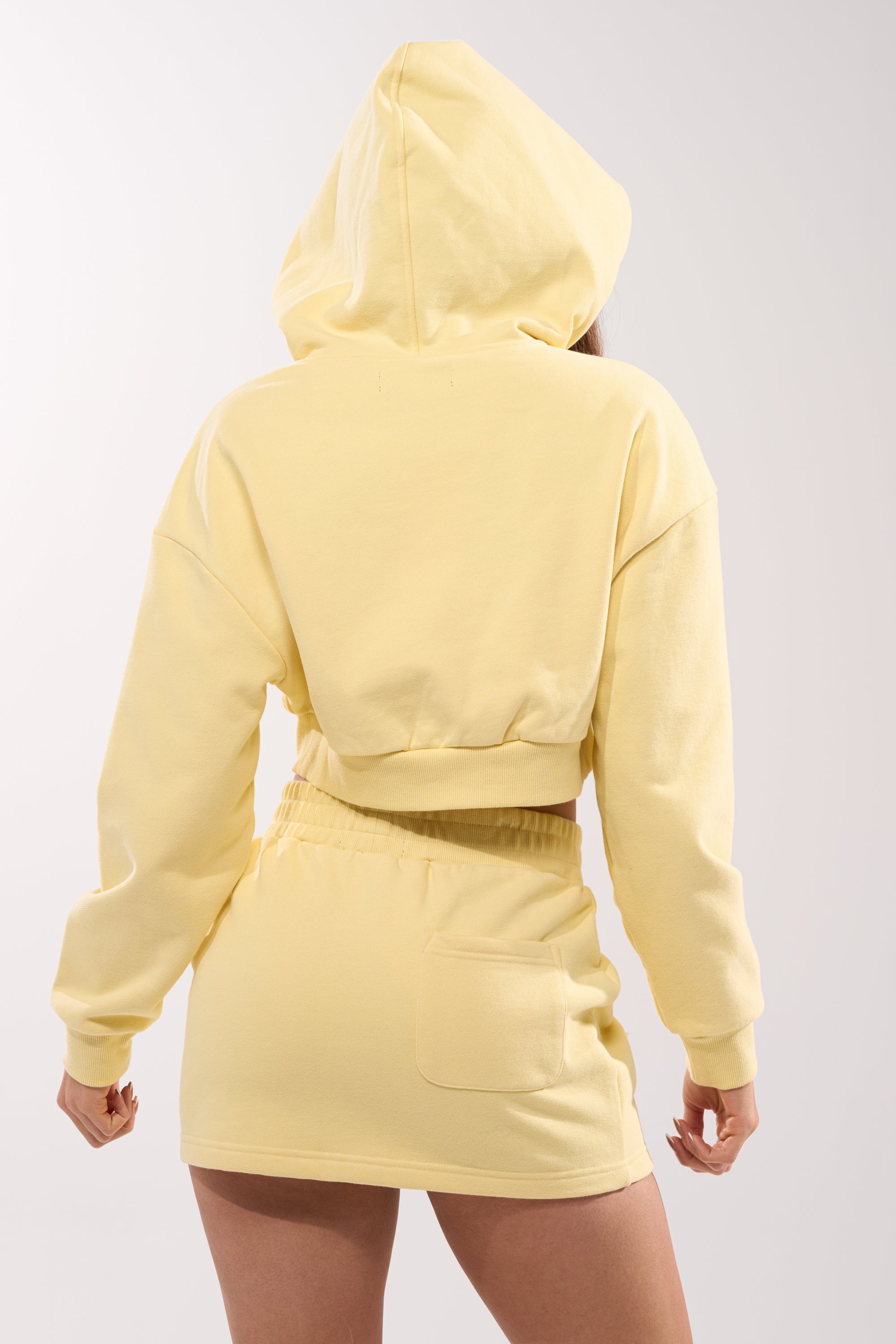 A person stands with their back to the camera, hood up, wearing the AKIRA Label AFTER HOURS SWEATSHIRT IN YELLOW and matching skirt with a right-side pocket, set against a plain light background. 5