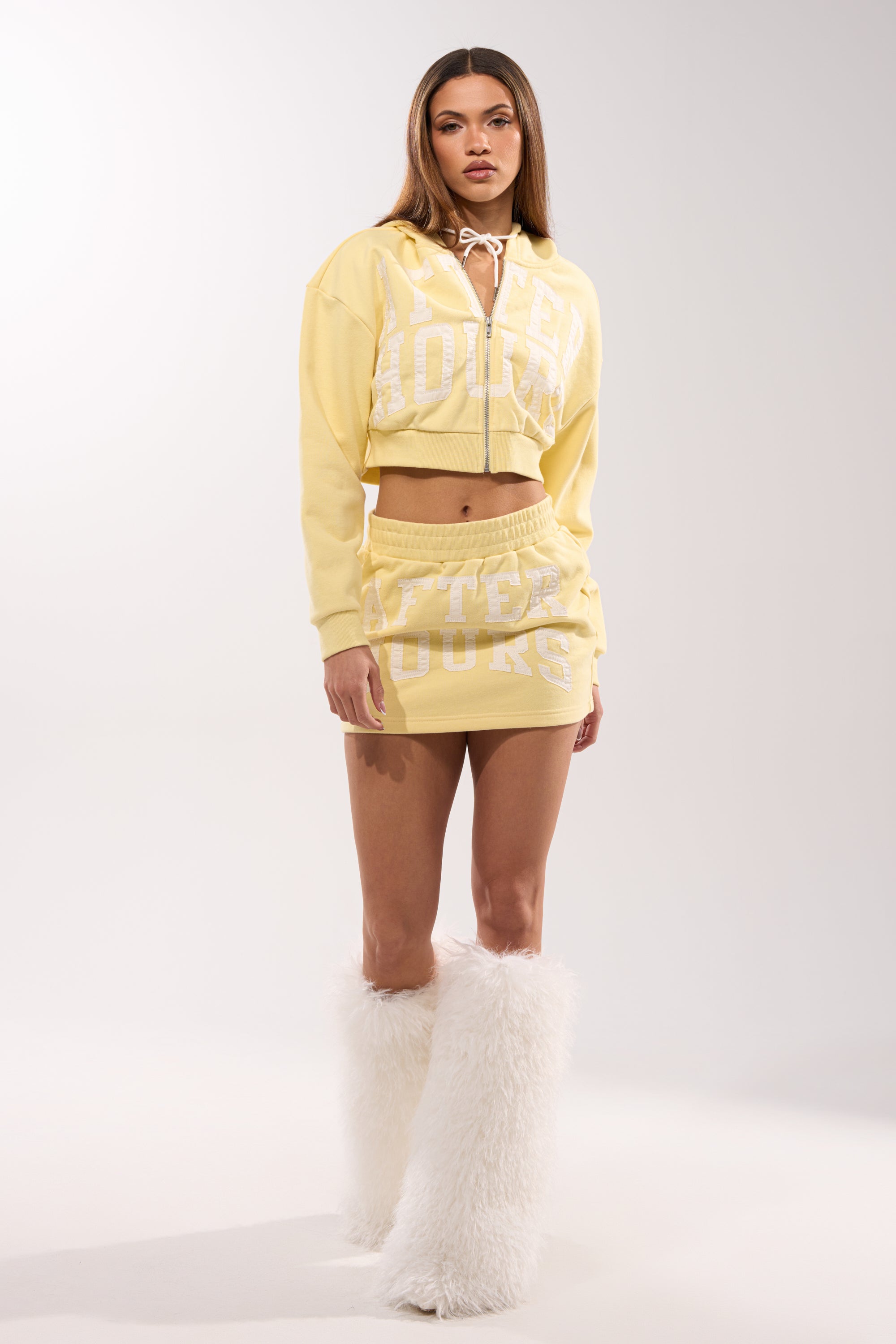 A woman with long hair and a neutral expression wears the AFTER HOURS SWEATSHIRT IN YELLOW, a matching mini skirt, and tall white boots against a plain background. 6