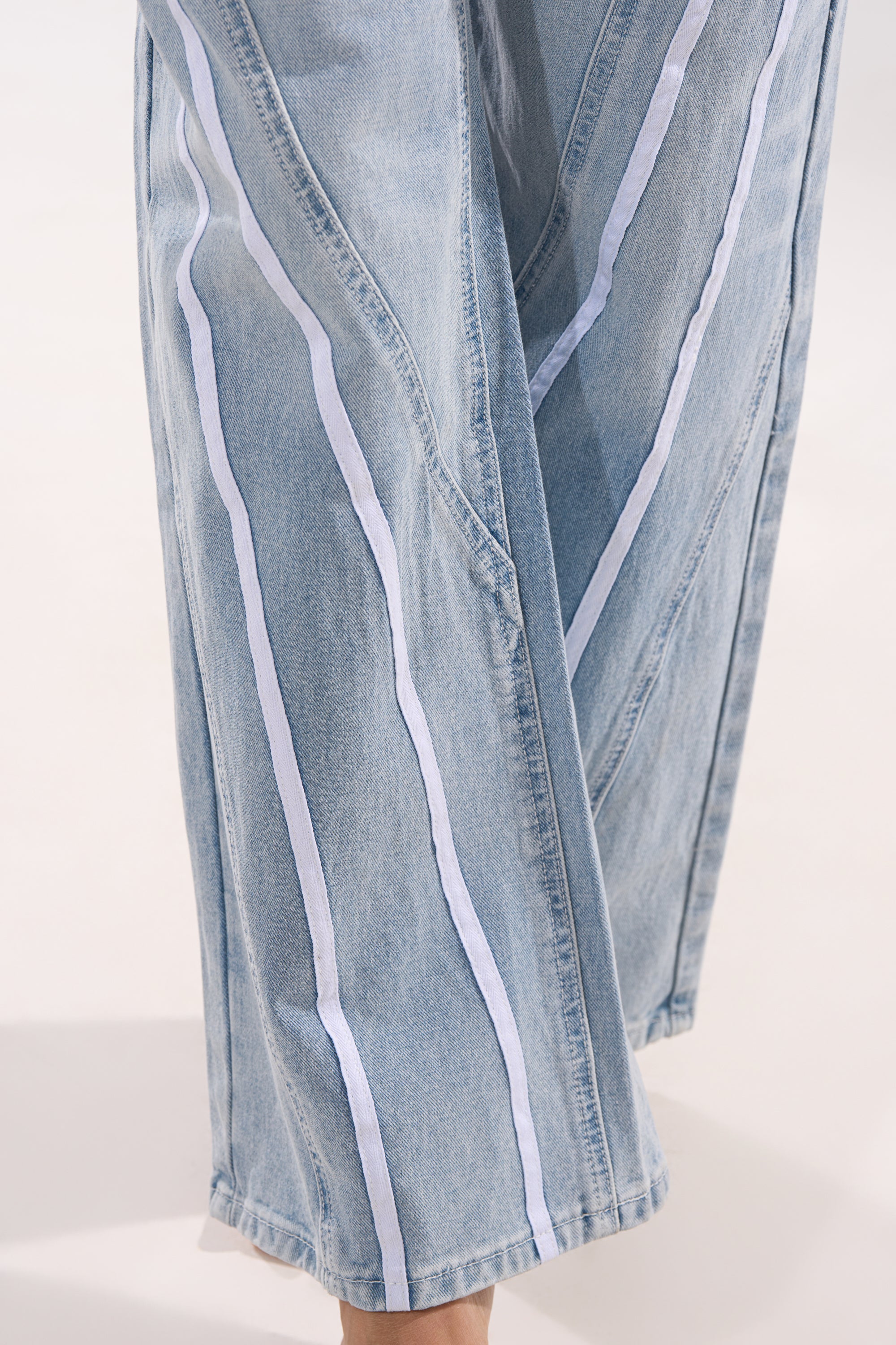 Close-up of the SHE IS A RUNNER DENIM TRACK PANT: light-wash, wide-leg jeans with vertical white stripes along the legs, displayed against a neutral background. 9