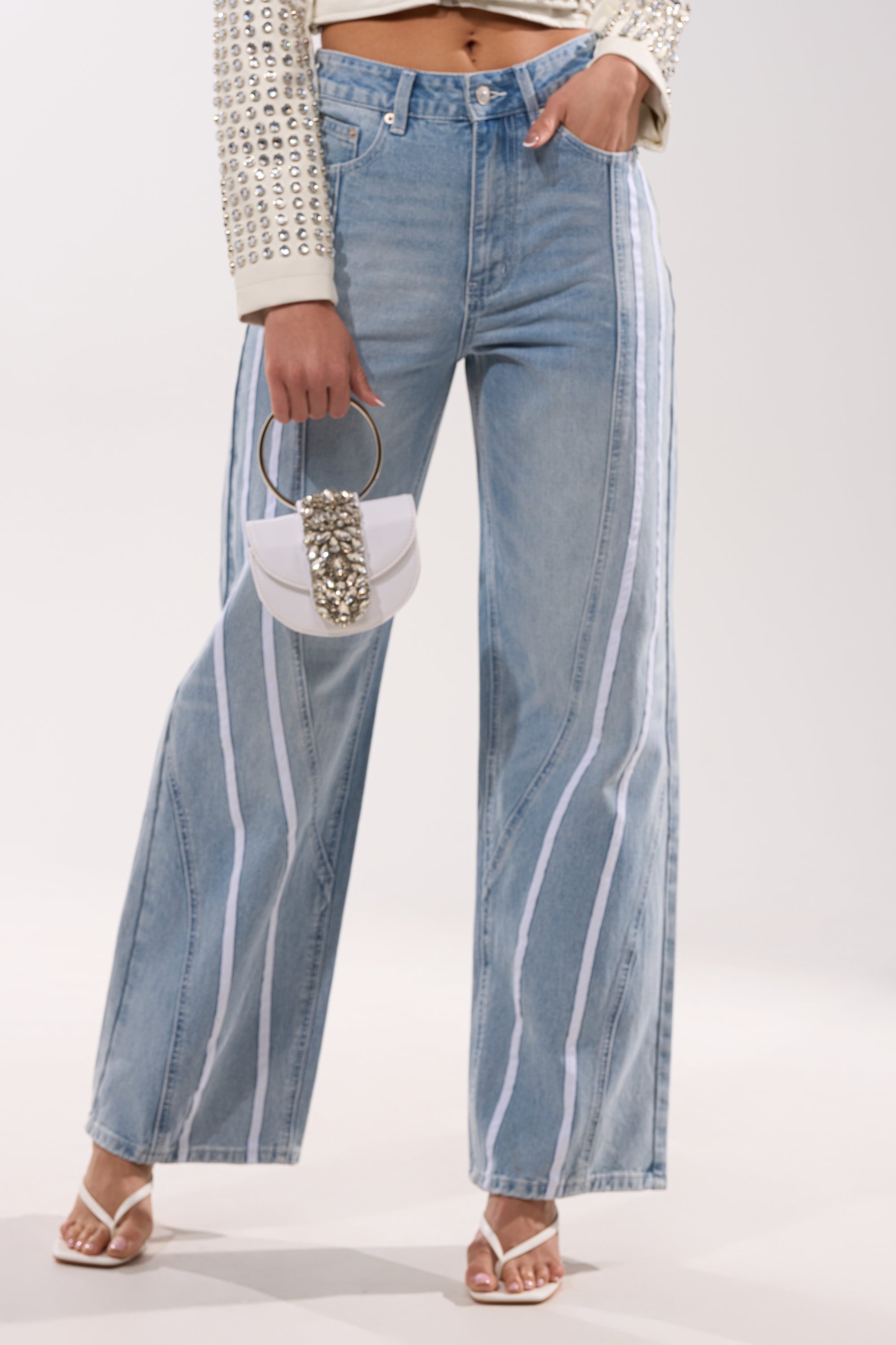A person wears the SHE IS A RUNNER DENIM TRACK PANT—high-waisted, wide-leg light-wash jeans with white vertical stripes—paired with a studded beige jacket, white heeled sandals, and a small metallic-detailed white purse. 1