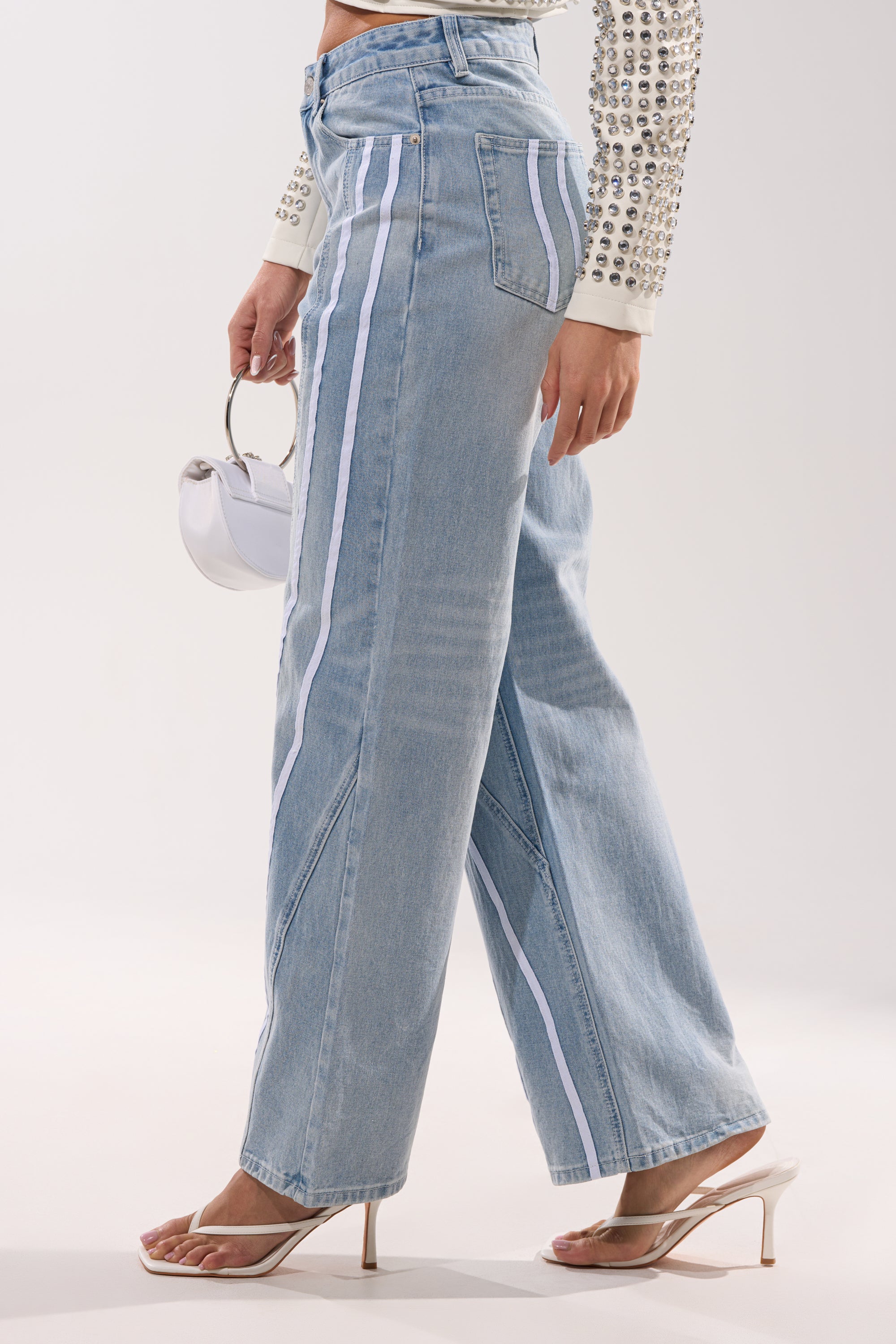 A person stands against a white background wearing the SHE IS A RUNNER DENIM TRACK PANT—light-wash wide-leg jeans with white side stripes—paired with a cream studded top, clear heeled sandals, and holding a small white handbag. 2