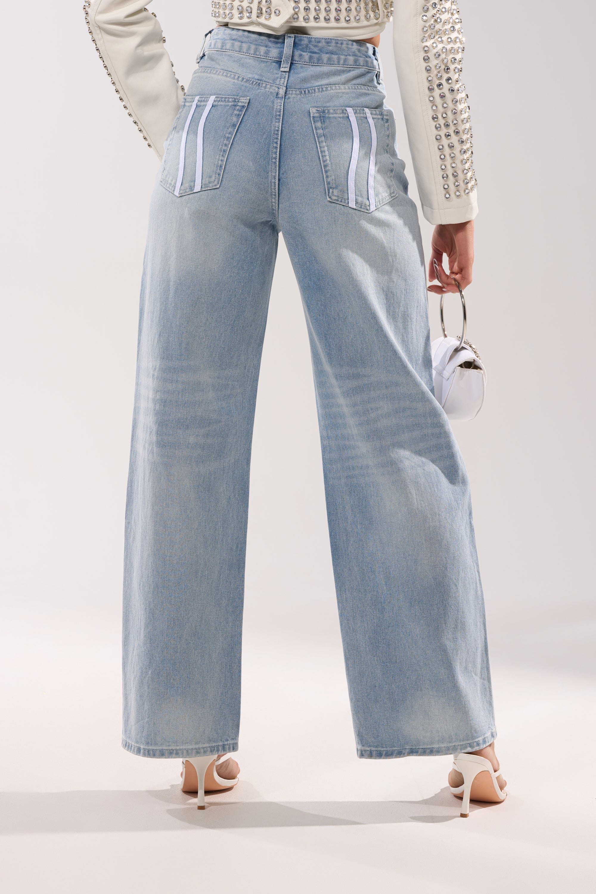 A person stands facing away, wearing the SHE IS A RUNNER DENIM TRACK PANT—high-rise wide-leg denim in light blue with white-striped back pockets—paired with a studded cream jacket, heeled sandals, and a small white handbag. 3
