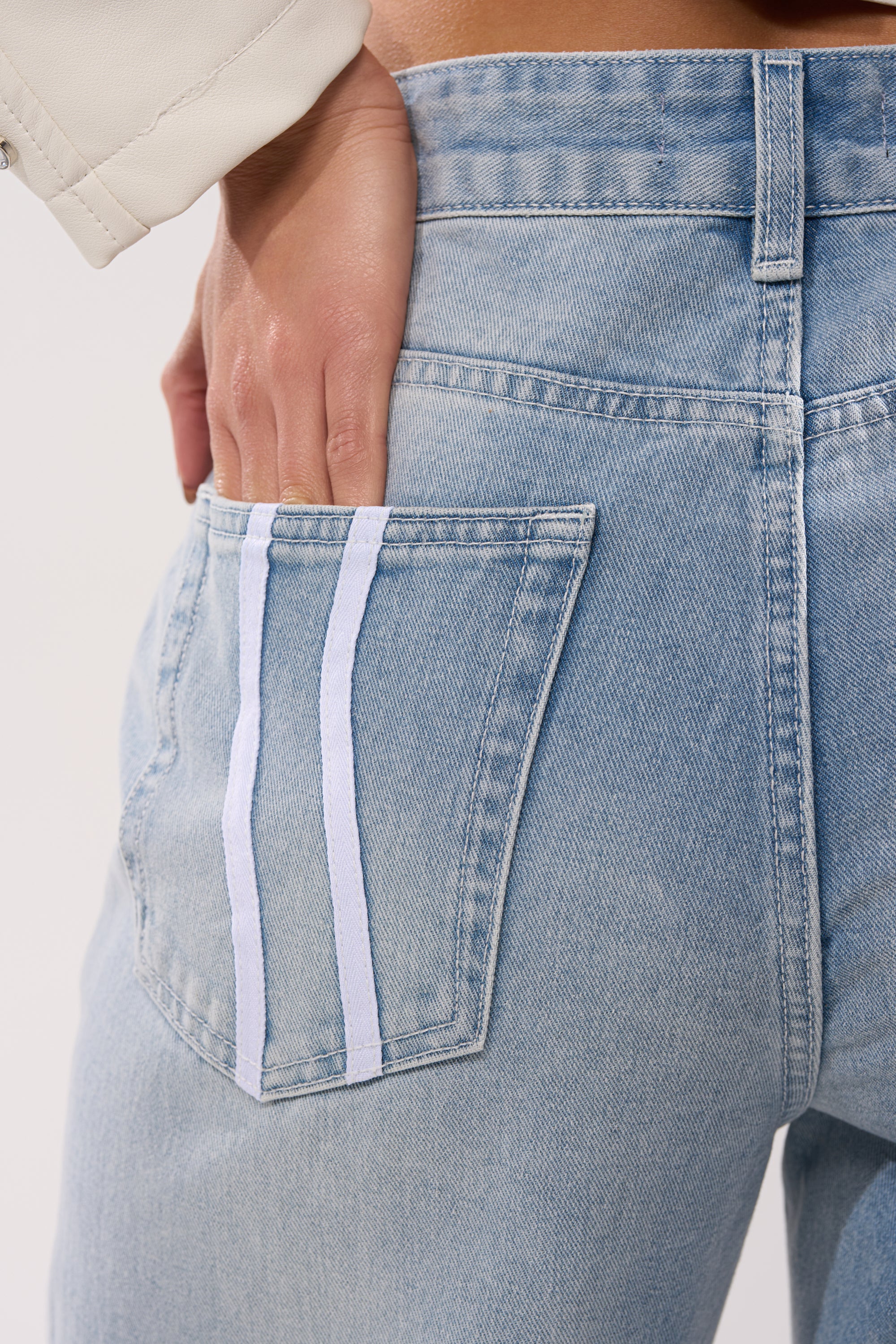 A person wearing the SHE IS A RUNNER DENIM TRACK PANT, featuring light-wash denim and two vertical white stripes on the back pocket, rests their left hand partially inside the pocket. 8