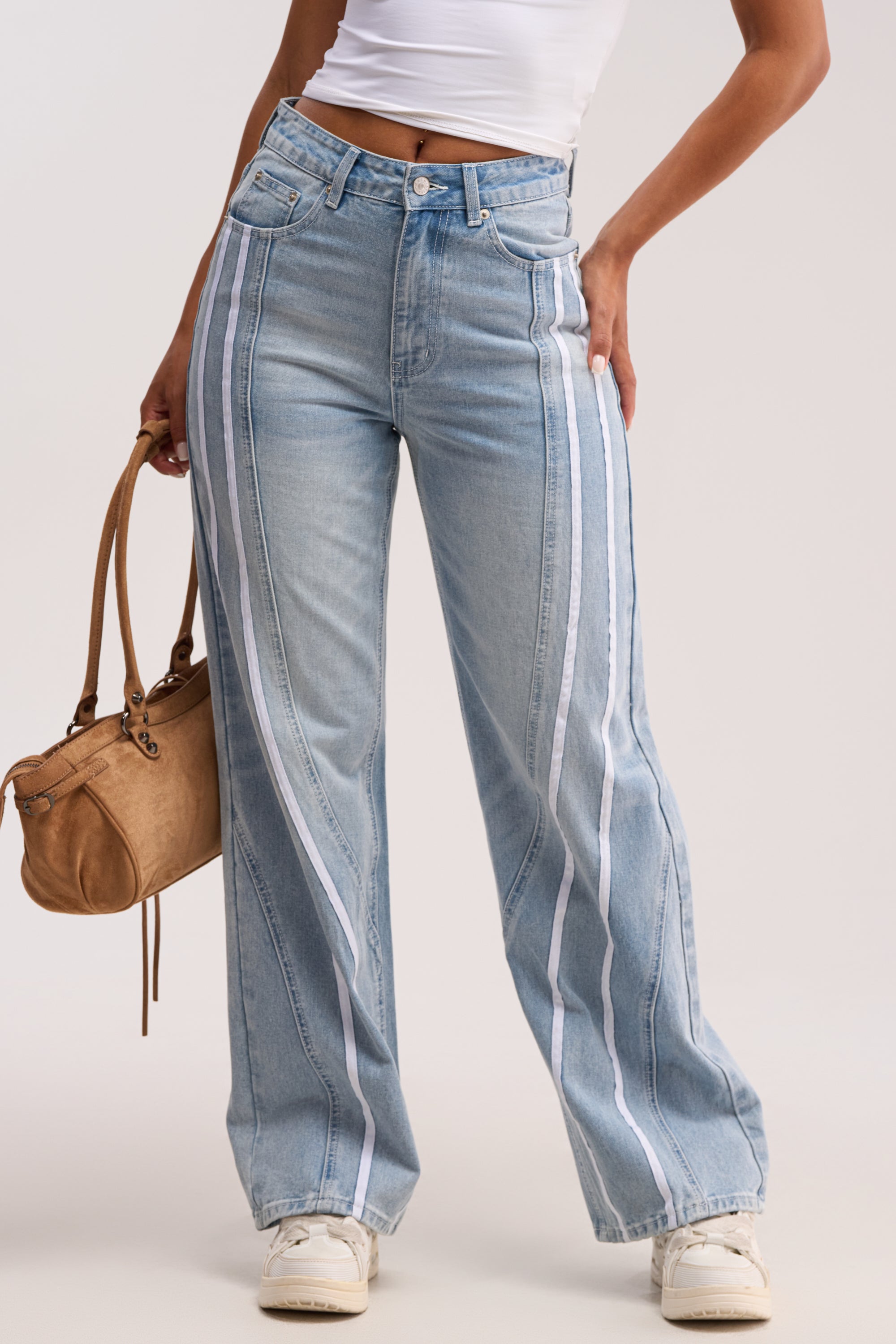 Wearing the SHE IS A RUNNER DENIM TRACK PANT—high-waisted, wide-leg jeans with double stripe side panels—paired with a white crop top, white sneakers, and a tan handbag against a plain, light backdrop.