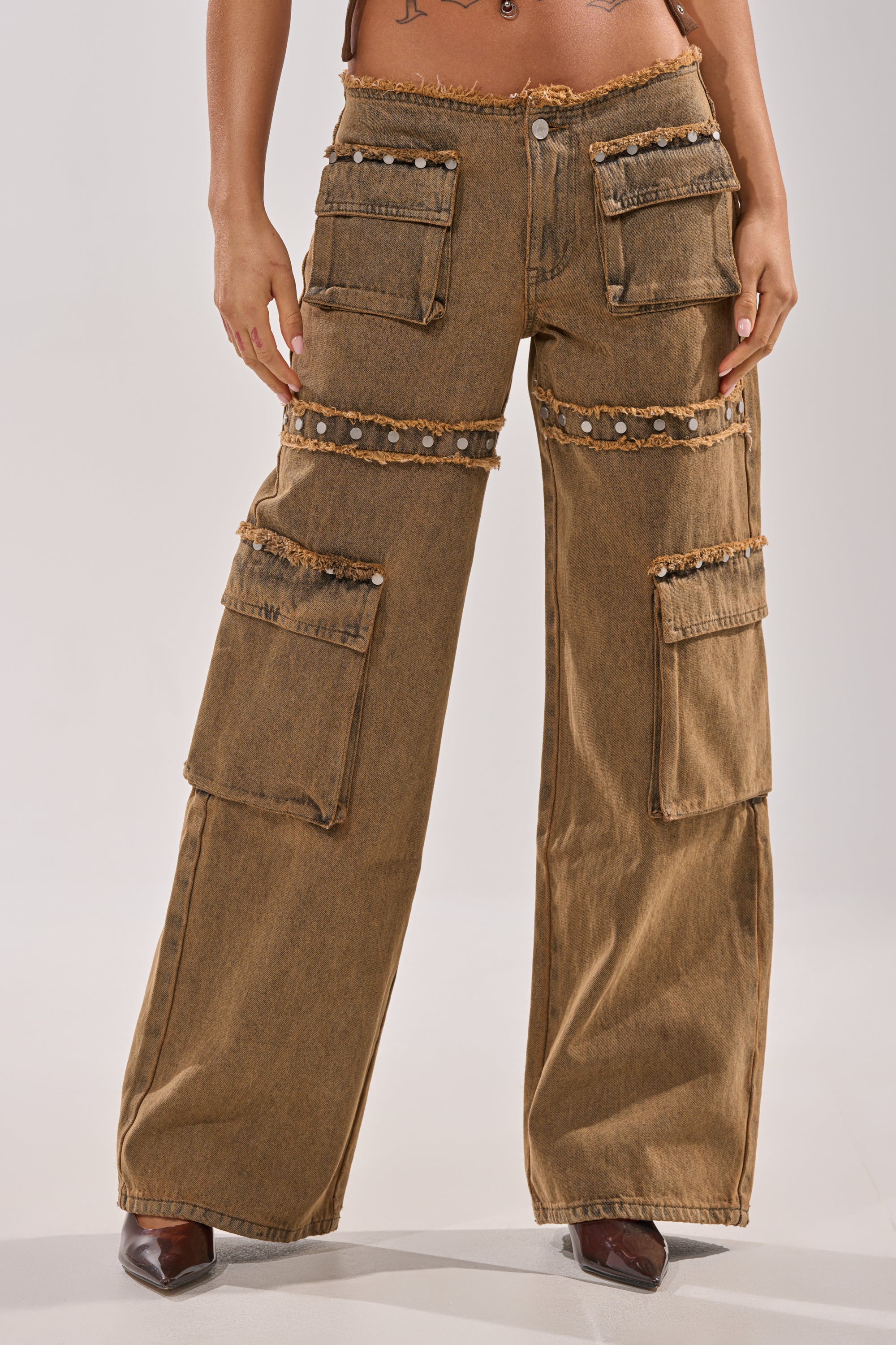 Someone wearing the OUT THE MUD DENIM JEANS—high-rise, wide-leg brown denim with cargo pockets, frayed edges, and decorative studs—paired with pointed brown shoes. 1