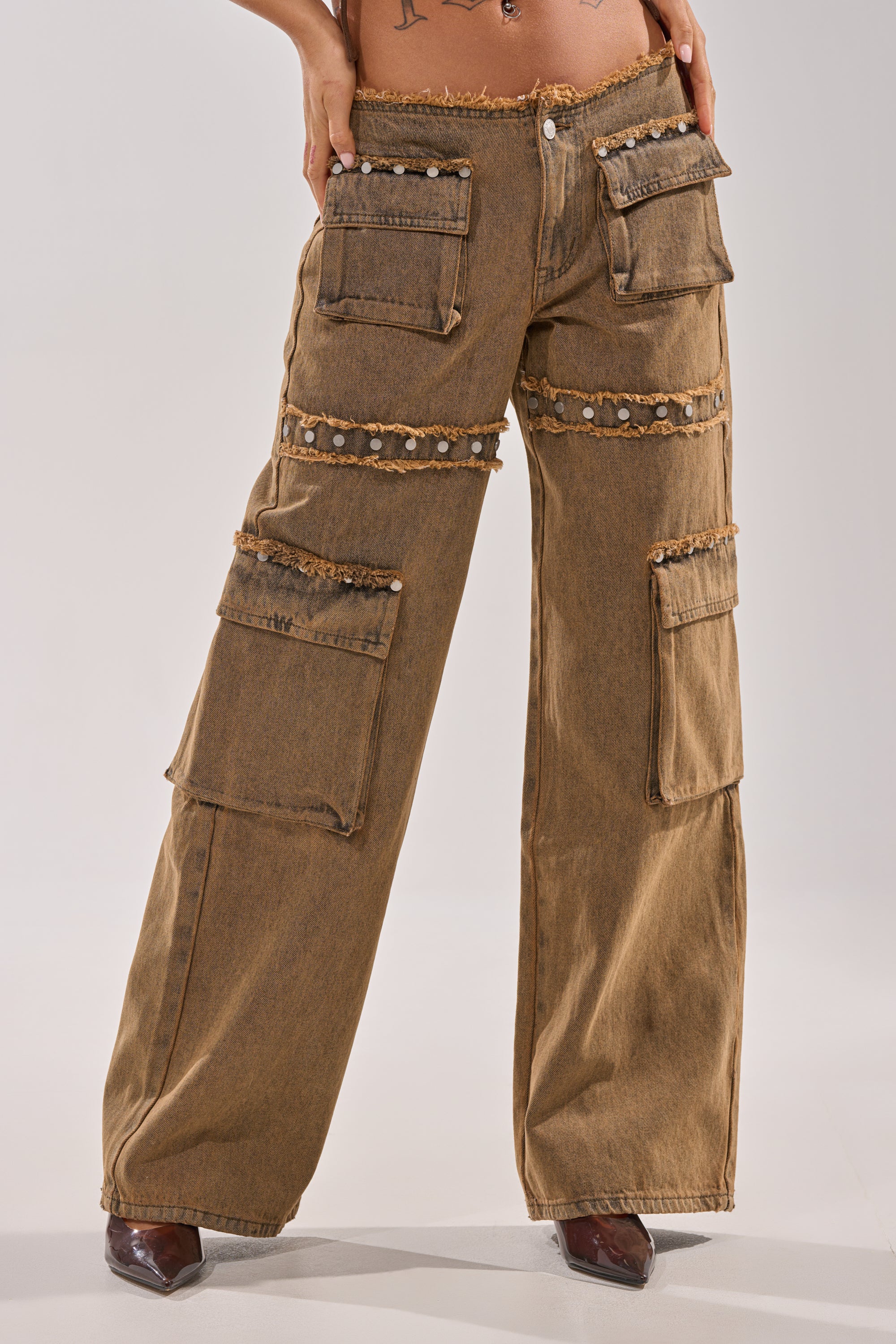 A person wears OUT THE MUD DENIM JEANS—high-rise, straight-leg brown denim with cargo pockets, frayed edges, and studs—paired with pointed brown shoes. Only the lower torso and legs are shown. 2