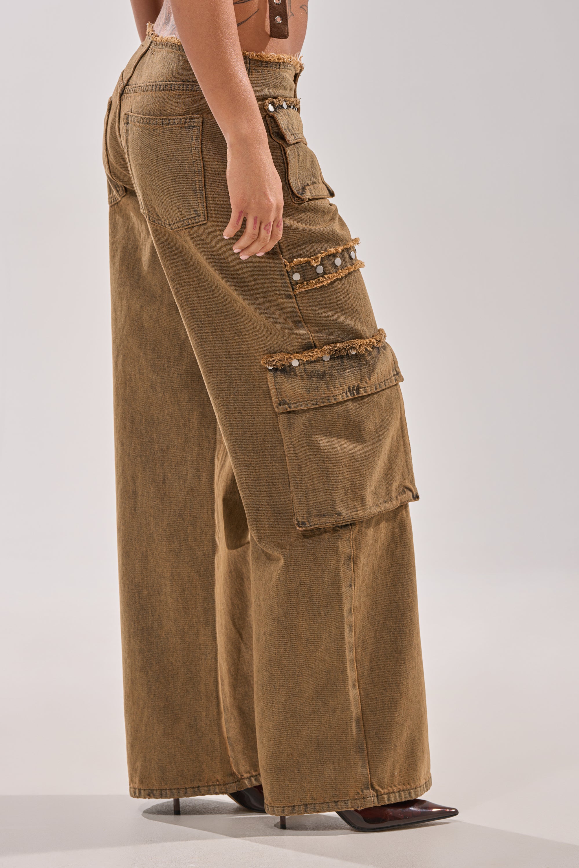 A person stands against a plain light background wearing OUT THE MUD DENIM JEANS—wide-leg brown cargo style with large front and side pockets, silver buttons, frayed edges—and pointed brown high-heeled shoes. 3