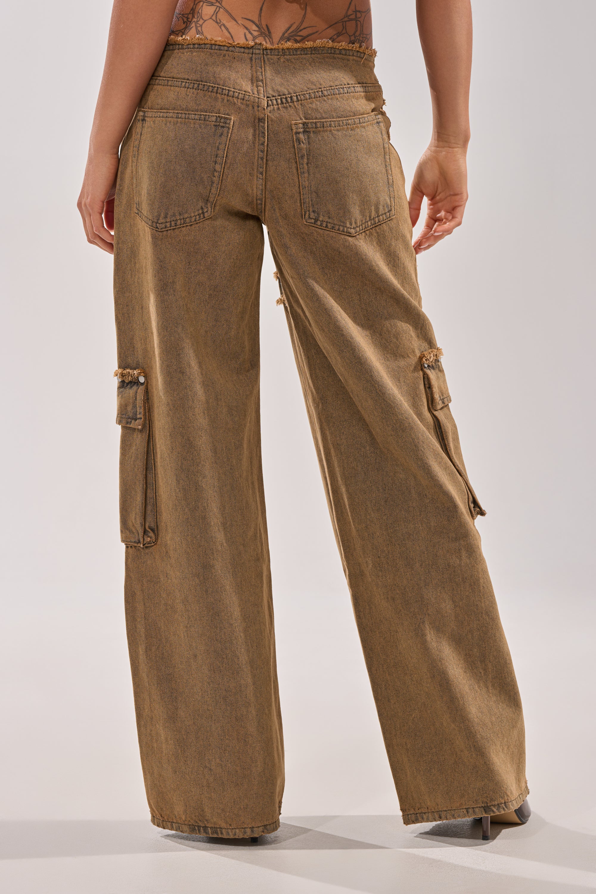 A person faces away, wearing OUT THE MUD DENIM JEANS—loose-fitting brown denim with a high-rise, straight-leg fit, cargo-style large back and side pockets, visible stitching, and partial upper back and arm in view. 4