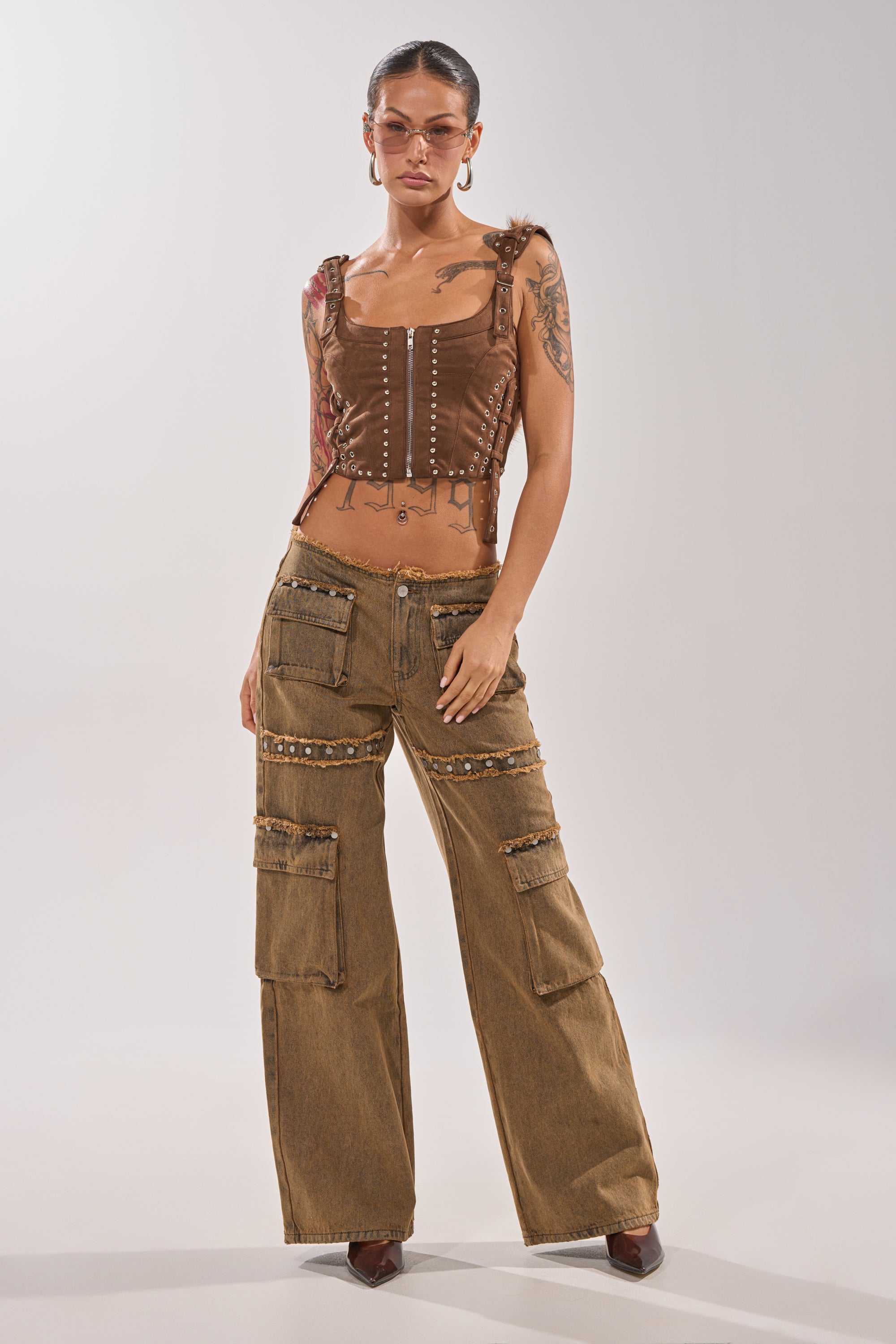A woman wearing OUT THE MUD DENIM JEANS with a brown corset-style top, brown pointed shoes, glasses, hoop earrings, and visible tattoos poses confidently against a plain background. 5