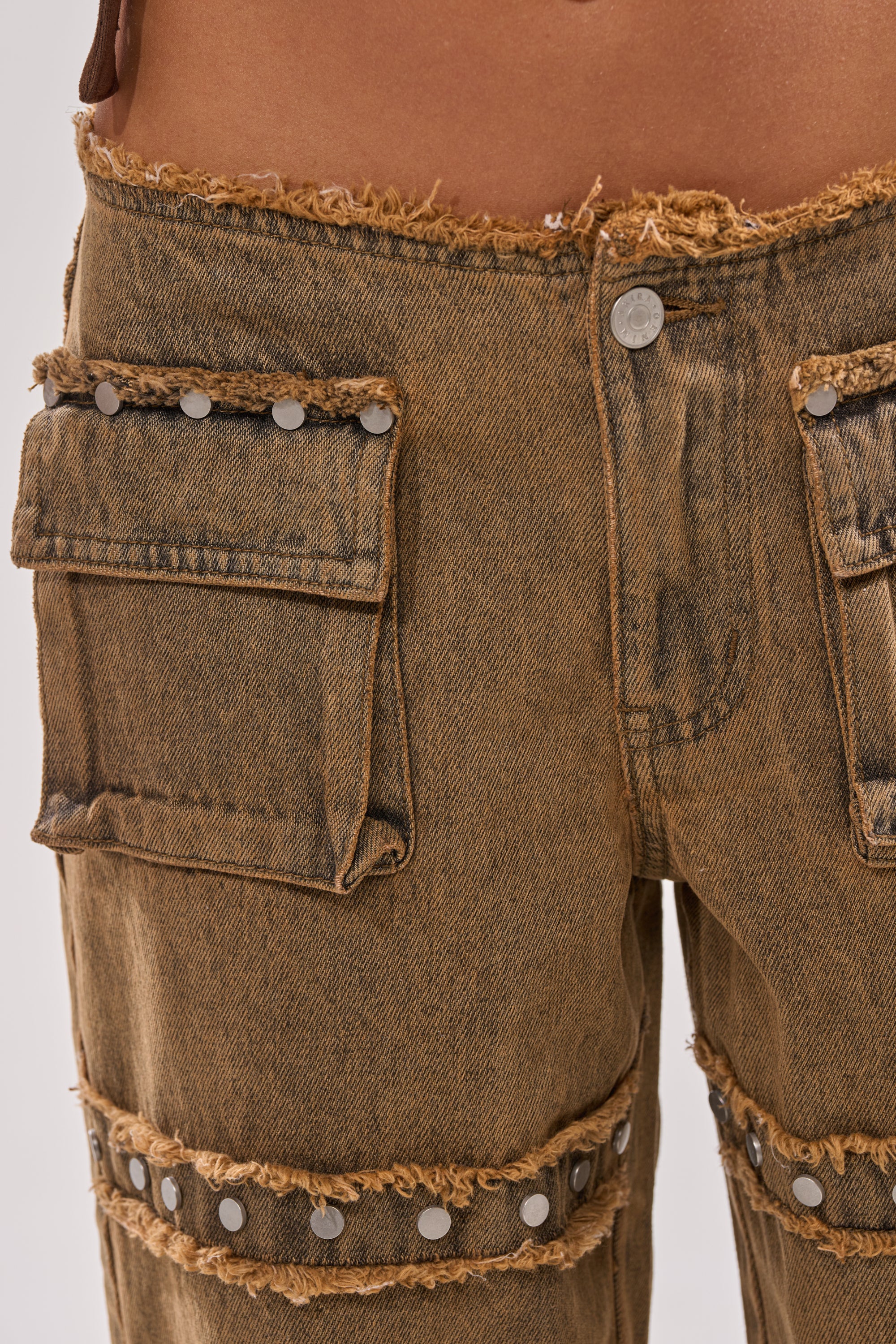 Close-up of a person wearing OUT THE MUD DENIM JEANS: high-rise, straight-leg brown denim with frayed hems, large cargo pockets with silver rivet details, a metal waistband button, and a worn, faded finish. 7