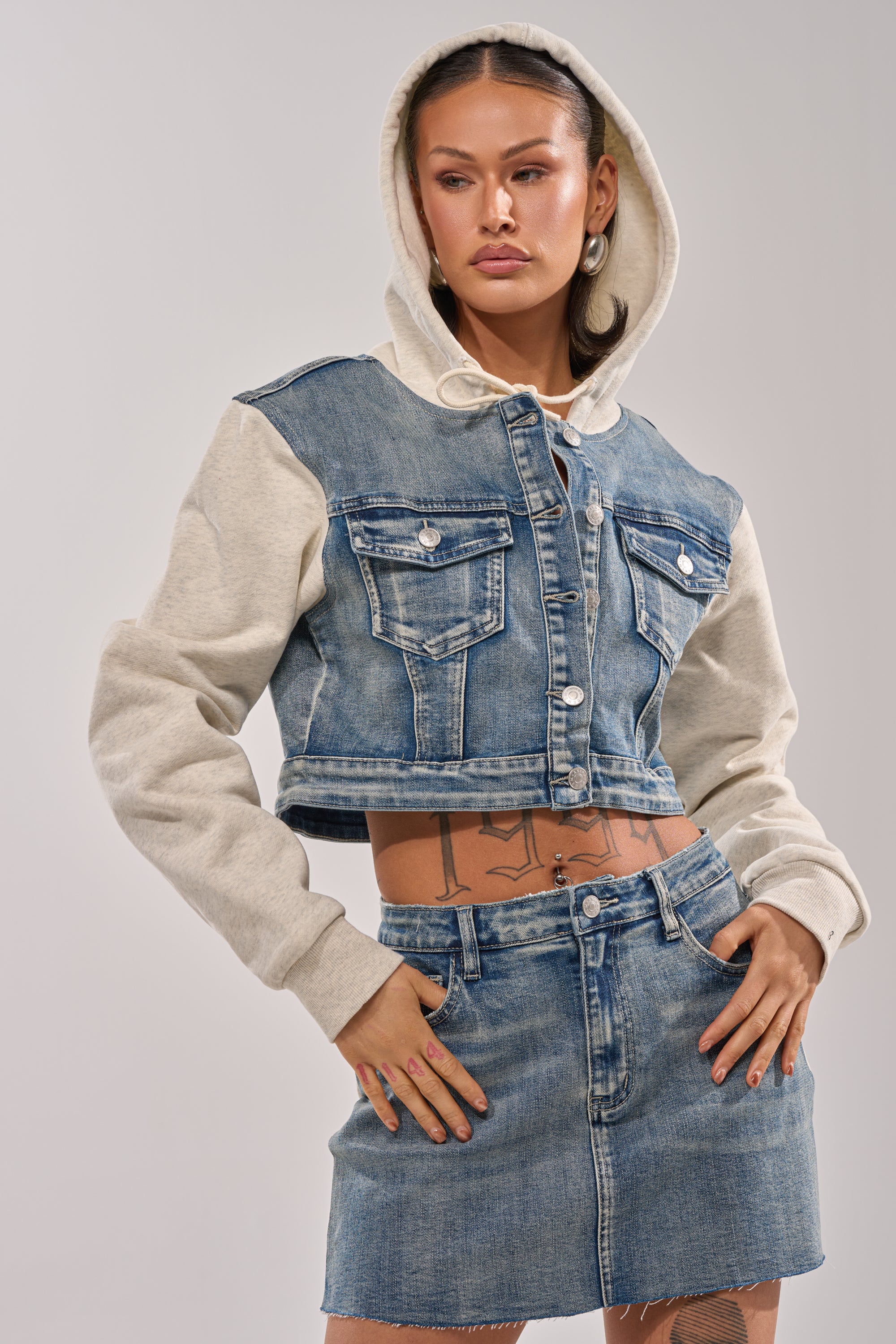A woman poses confidently in the AFTER HOURS DENIM JACKET, featuring a fleece hood and light gray sleeves, paired with a matching denim mini skirt. She accessorizes with hoop earrings and shows off her tattoos.