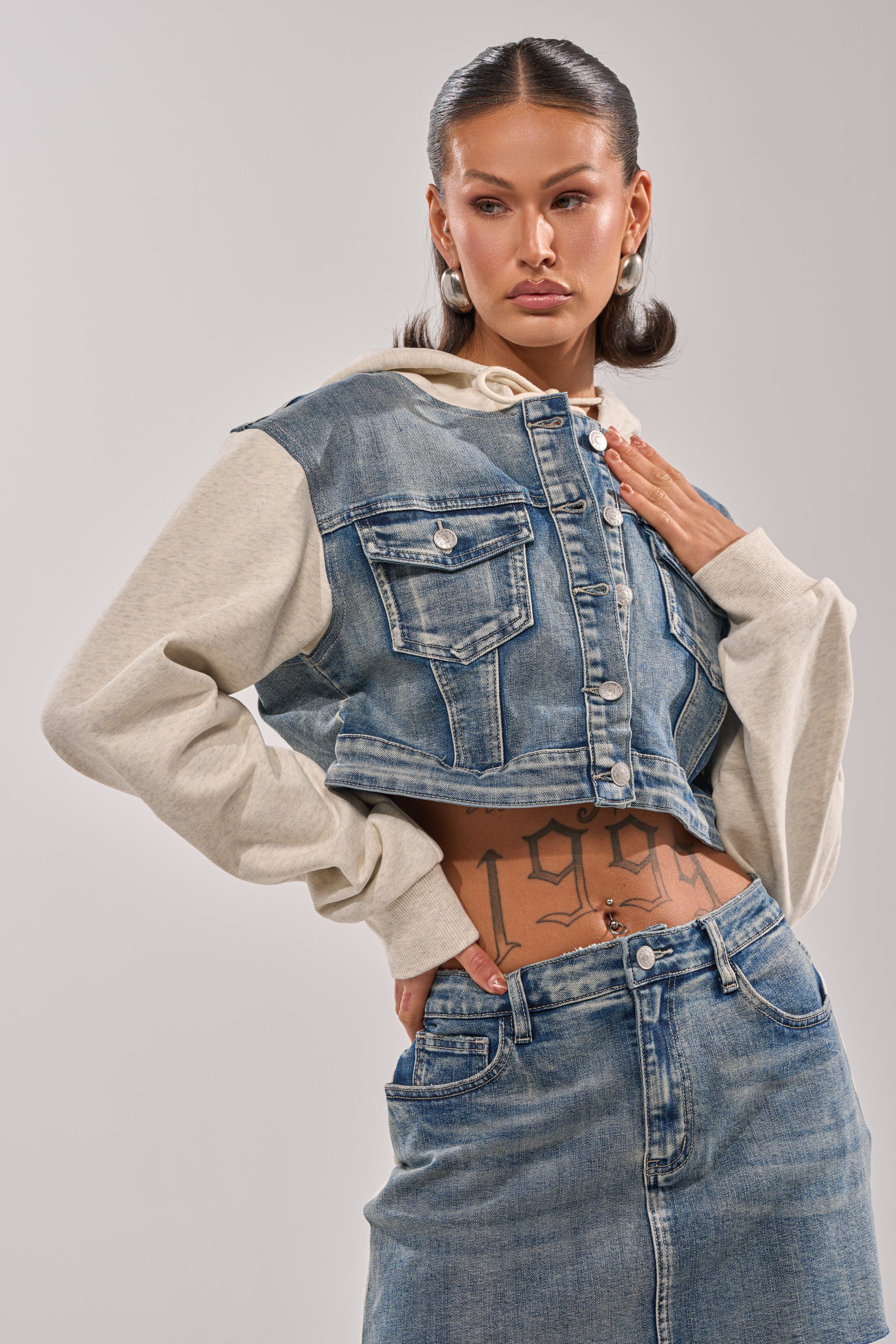 A woman poses confidently in the AFTER HOURS DENIM JACKET, featuring gray sleeves and a fleece hood, styled with a denim skirt. She wears pearl earrings, has slicked-back hair, and shows off a visible "1999" tattoo on her stomach.
