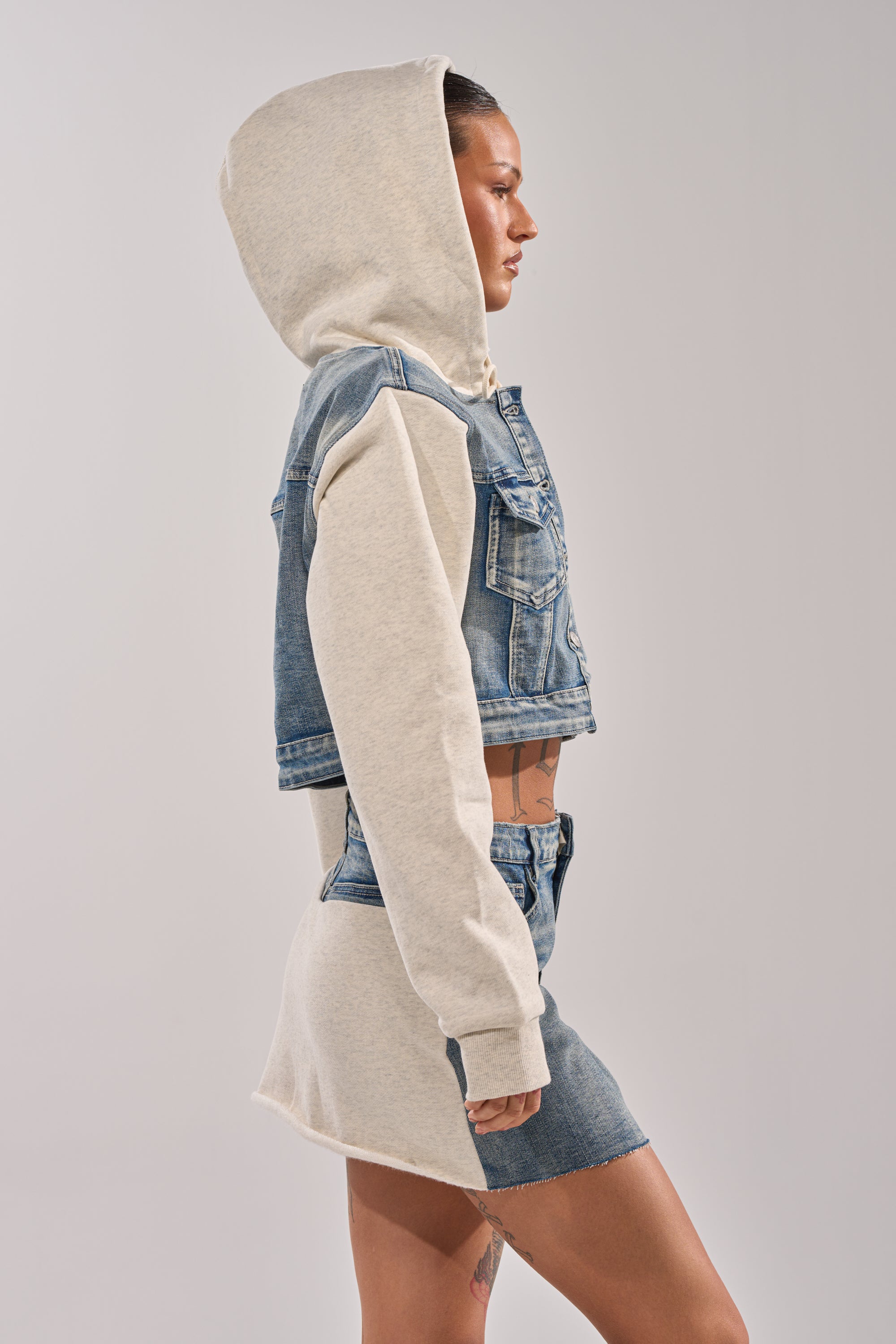 A woman in profile wears the AFTER HOURS DENIM JACKET—cropped with denim and light gray hoodie sleeves—paired with a matching two-tone denim and gray mini skirt, standing against a plain light background.
