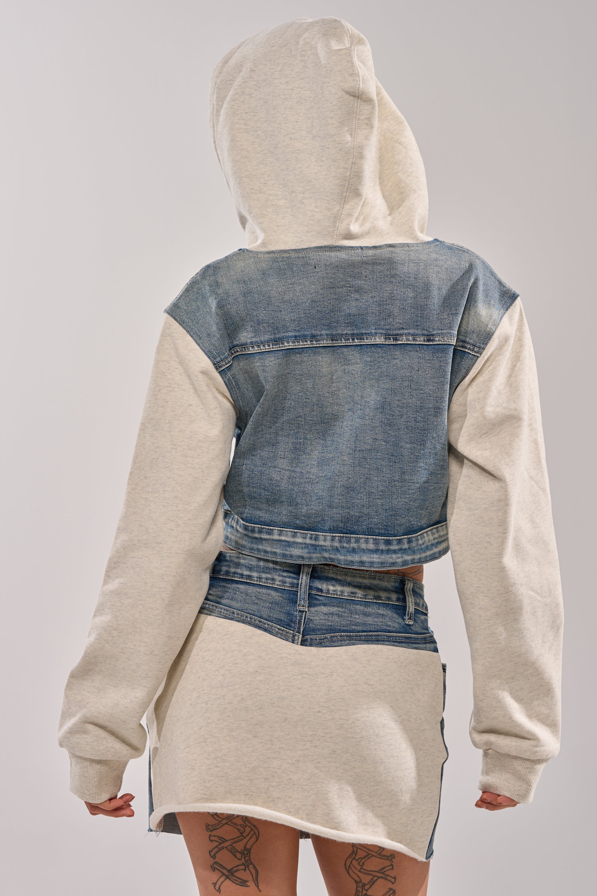 A person stands facing away, wearing the AFTER HOURS DENIM JACKET over a light gray hoodie and matching skirt. Their hands rest by their sides, revealing tattoos on the backs of their thighs.