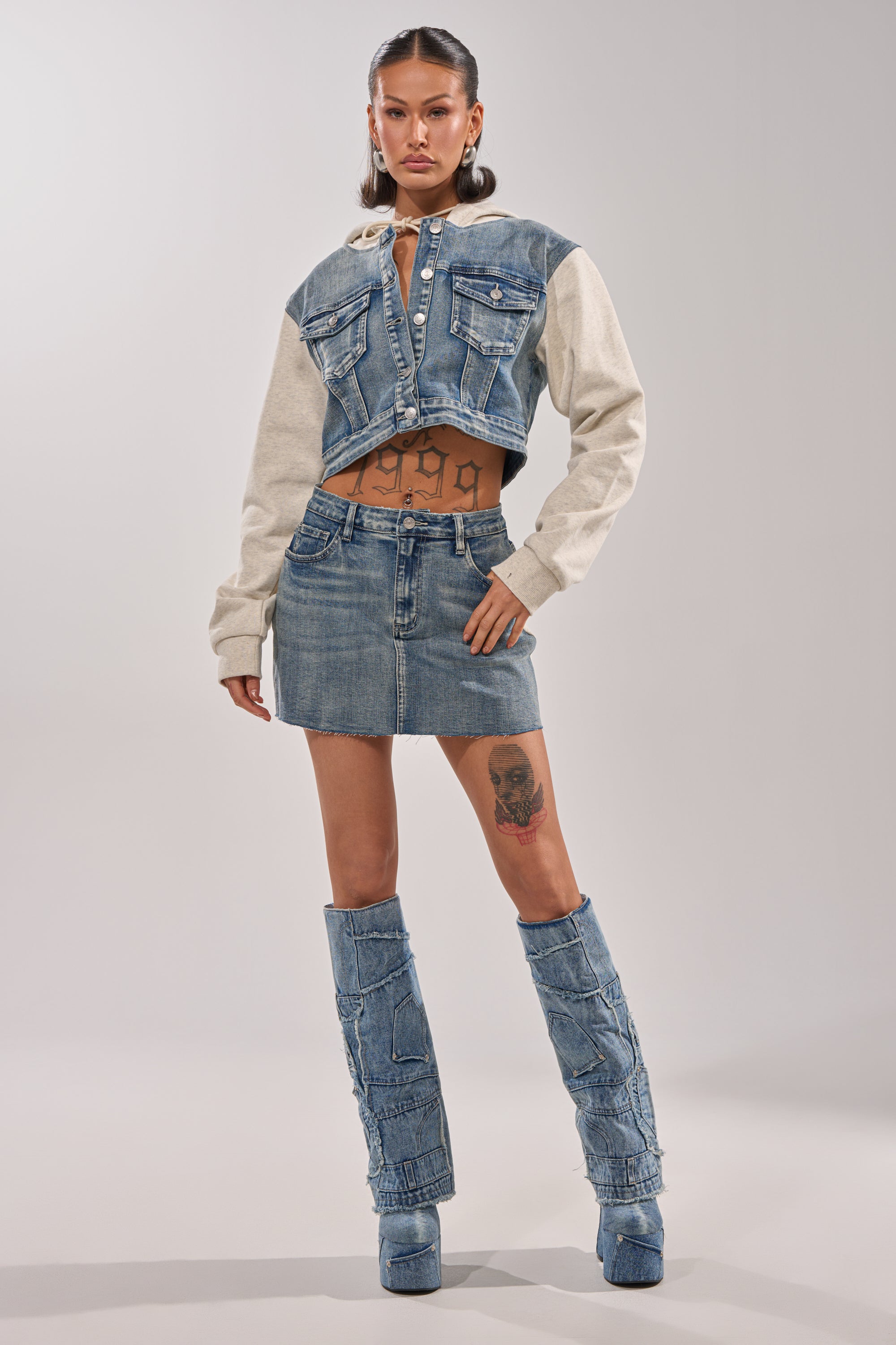 A woman models the AFTER HOURS DENIM JACKET, featuring a cropped fit with beige sleeves. She pairs it with a matching denim mini skirt and high boots, displaying tattoos on her torso and thigh against a plain light backdrop.