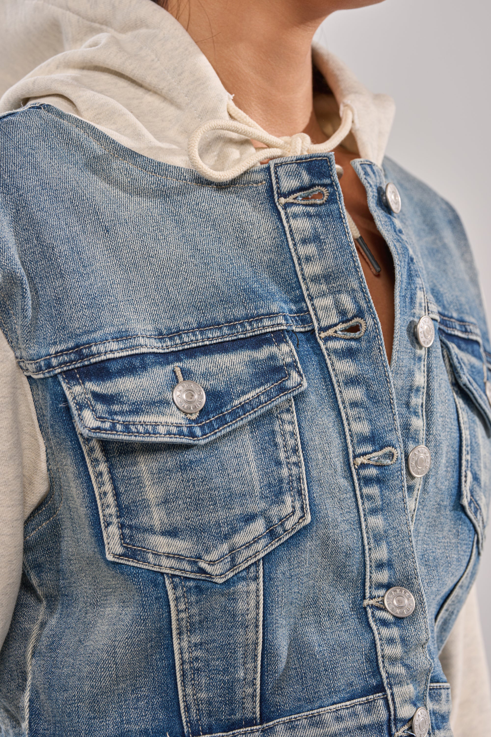 A person wears the AFTER HOURS DENIM JACKET, a cropped denim style with silver buttons and chest pockets, layered over a light hoodie. Only the upper torso and part of their face are visible.