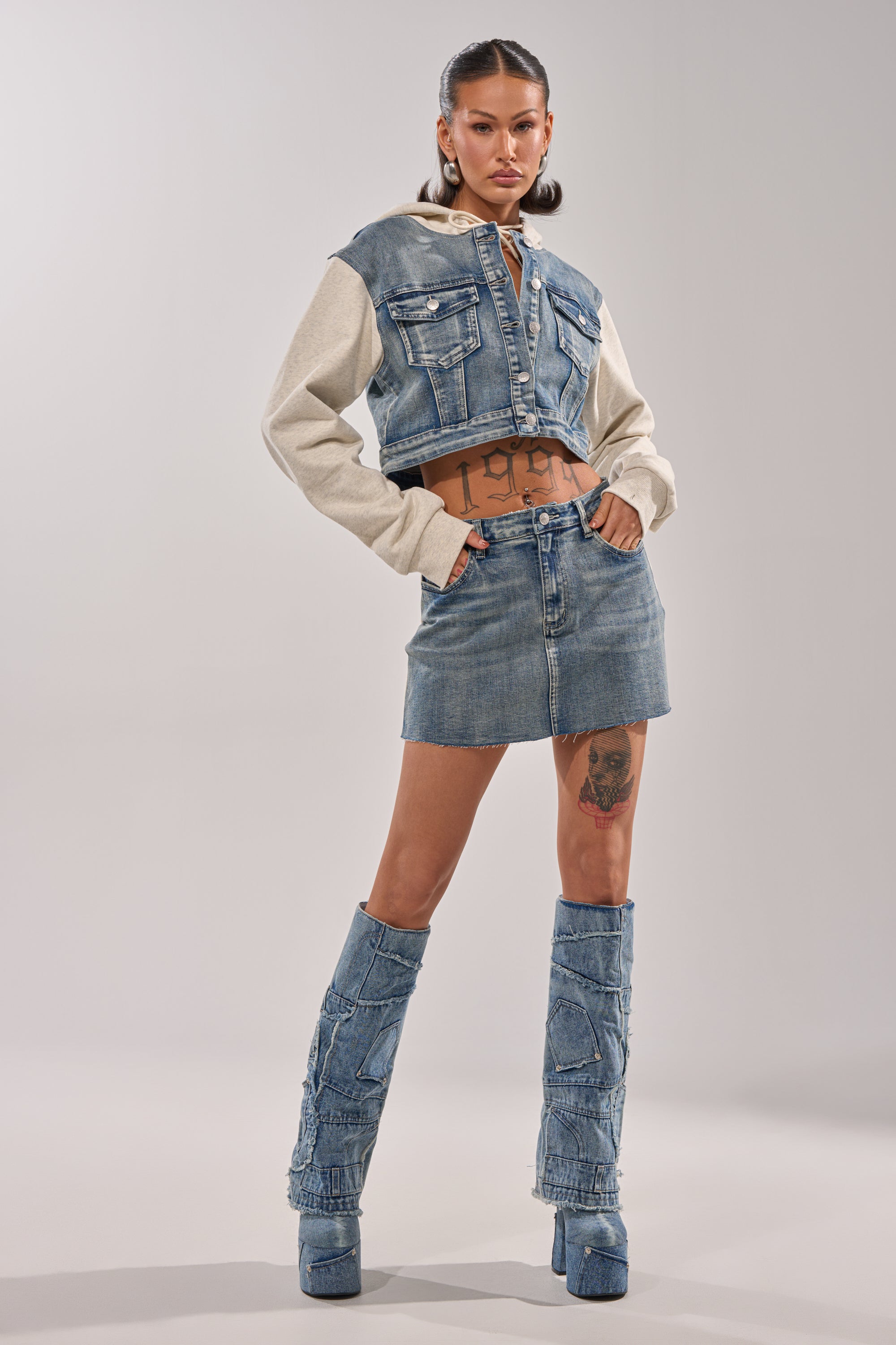 A woman wears the AFTER HOURS DENIM MINI SKIRT with a light denim jacket, beige sleeves, and knee-high denim boots. Her two ponytails and visible tattoos on her stomach and thigh complete her confident, bold style. 0