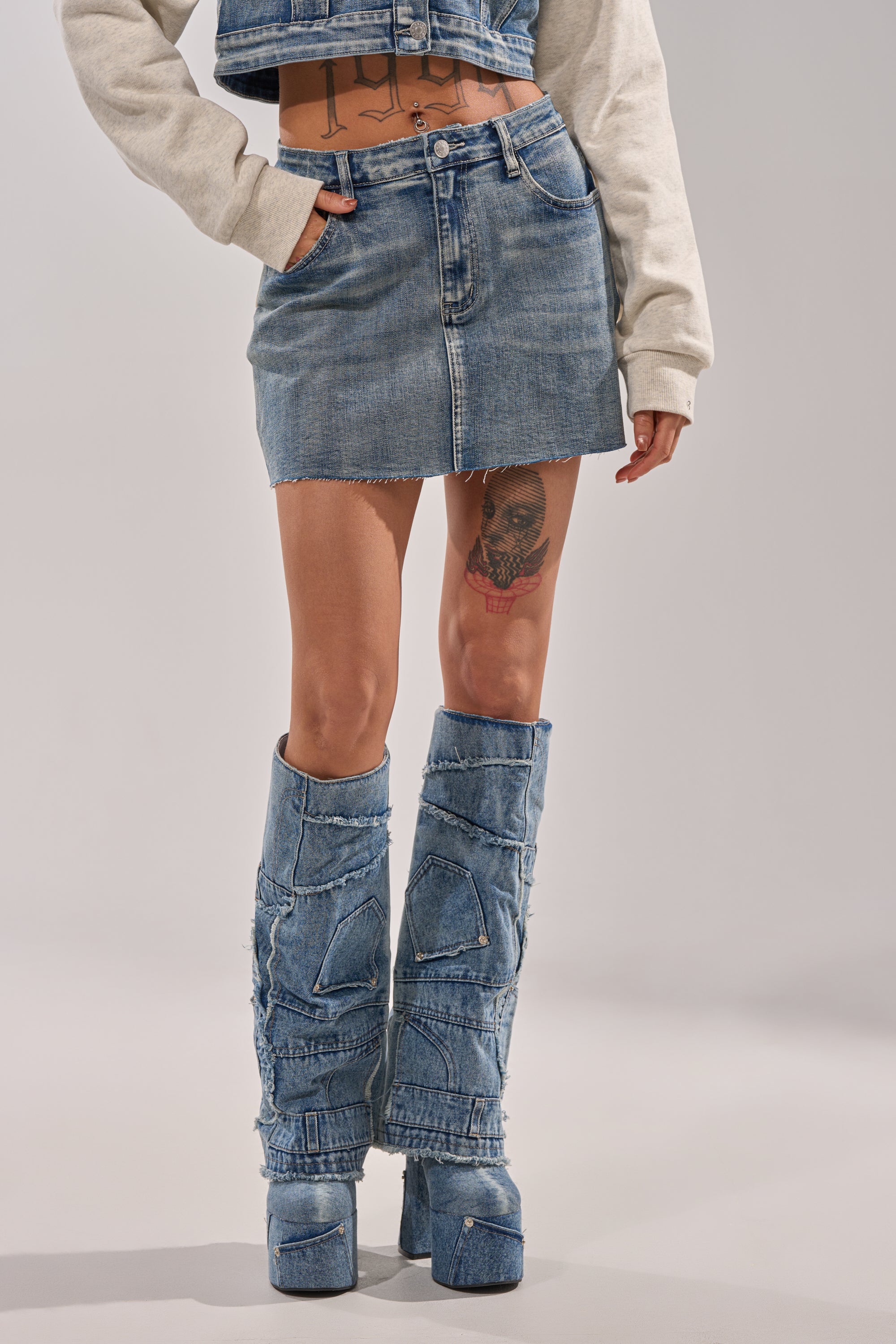 Wearing the AFTER HOURS DENIM MINI SKIRT with matching patchwork platform boots and a cropped jacket, a hand slips into a pocket, revealing tattoos on the stomach and thigh against a neutral background. 1