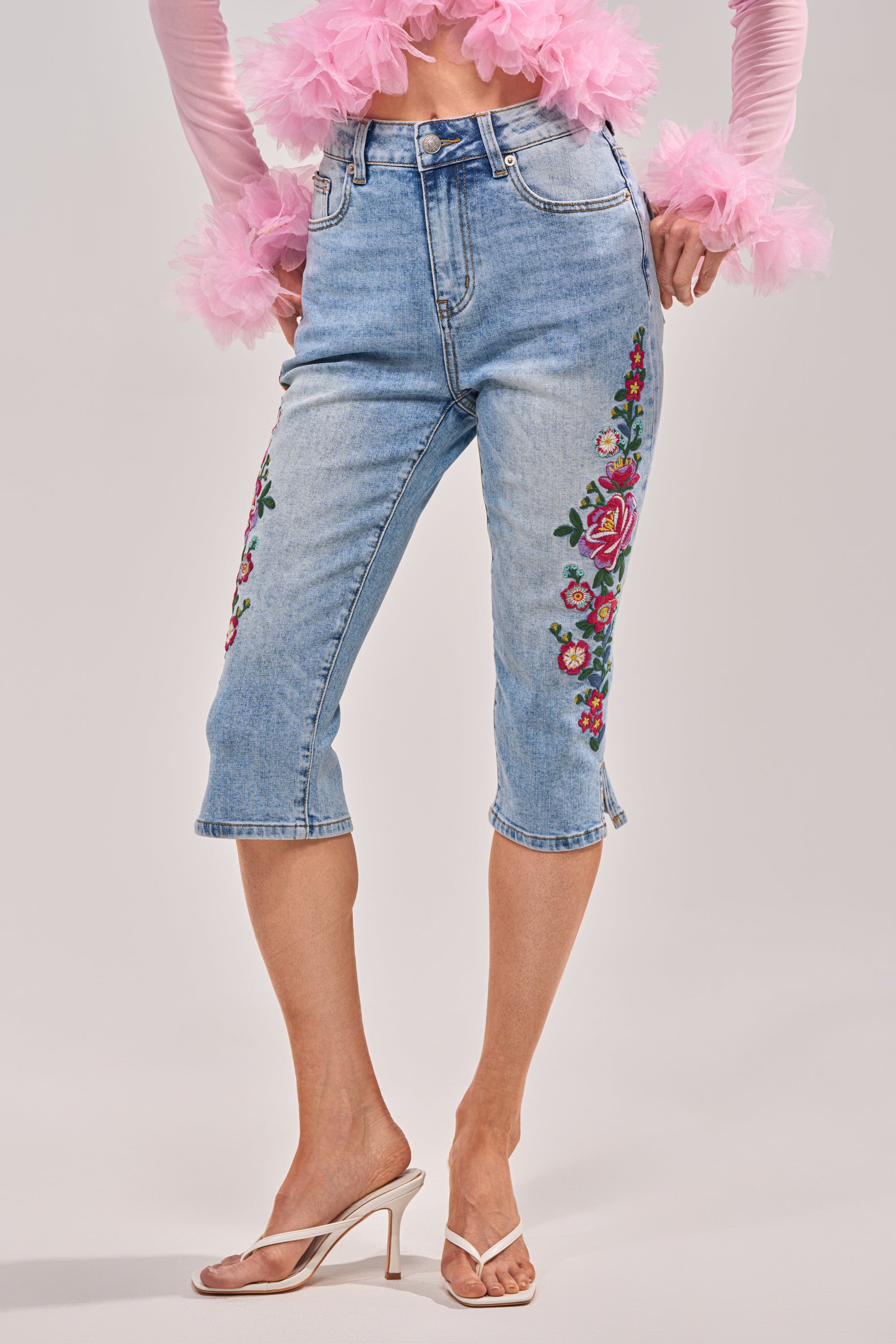 Someone wears the GIVE ME FLOWERS FLORAL CAPRI—floral embroidered, cropped denim capris—paired with clear strap high-heeled sandals and a sheer pink ruffled top, set against a plain light background. 0