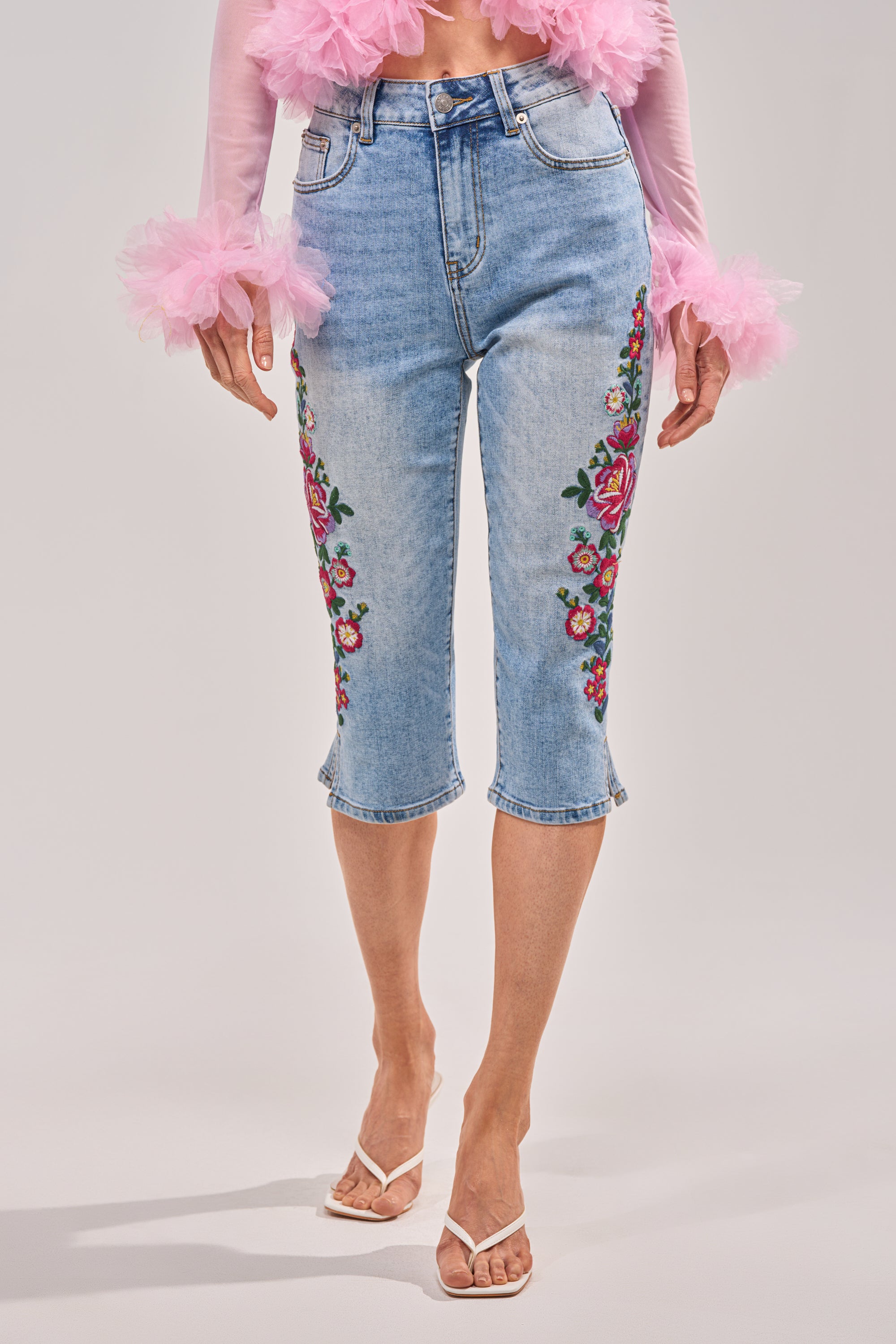 Wearing the GIVE ME FLOWERS FLORAL CAPRI, a pink sheer top with ruffled cuffs, and white heeled sandals, a person stands against a plain background. 1