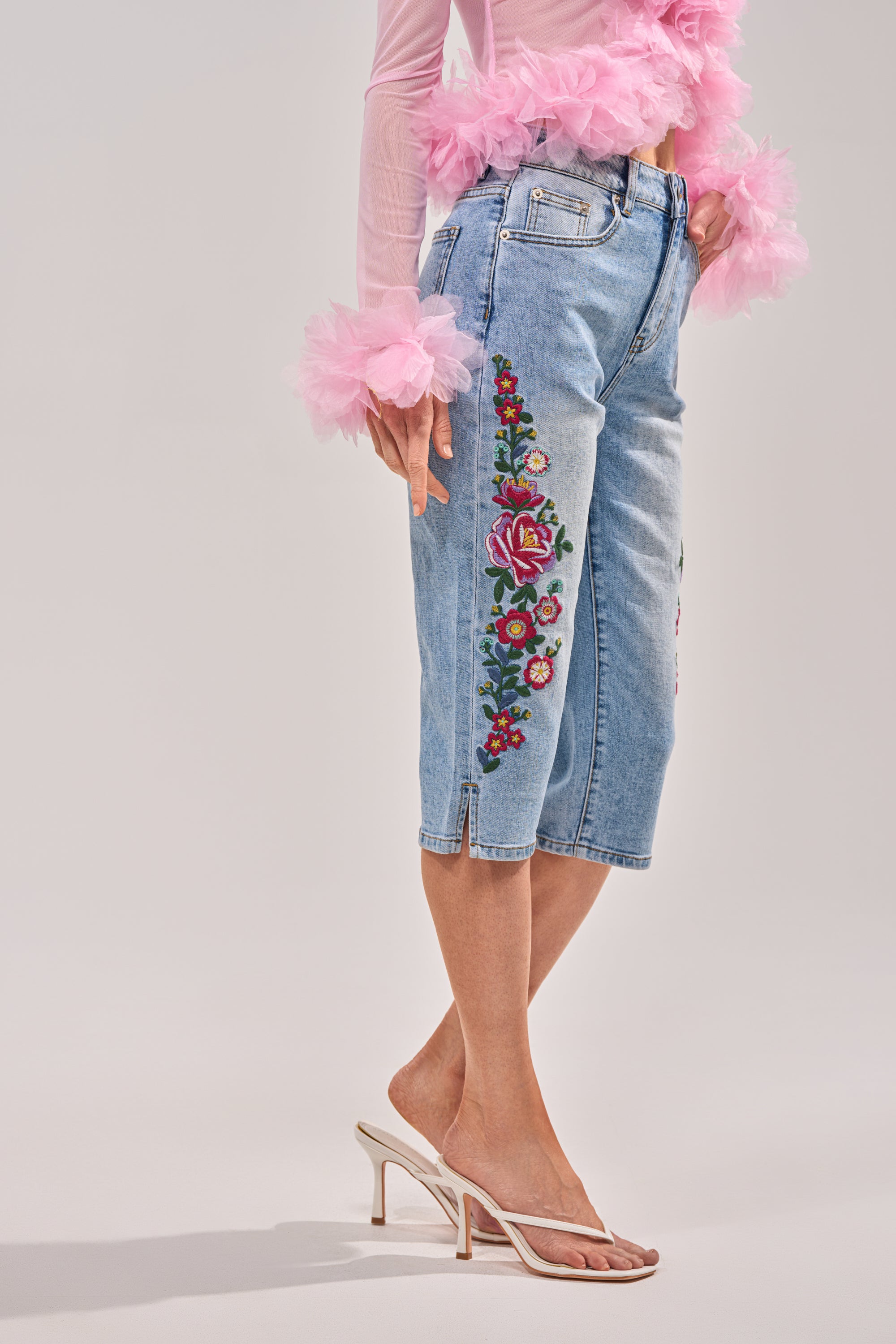 A woman models the GIVE ME FLOWERS FLORAL CAPRI with colorful side embroidery, paired with a sheer pink top with tulle cuffs and white heeled sandals, standing against a plain background. 2