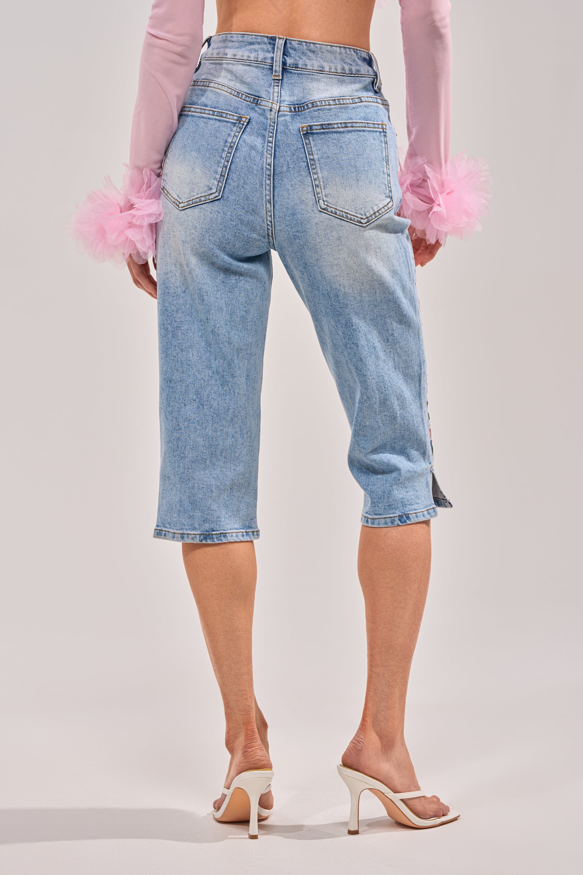 A person stands against a plain background, shown from the back, wearing GIVE ME FLOWERS FLORAL CAPRI pants in light blue, paired with a long-sleeve pink top featuring tulle ruffle cuffs and white high-heeled sandals. 3